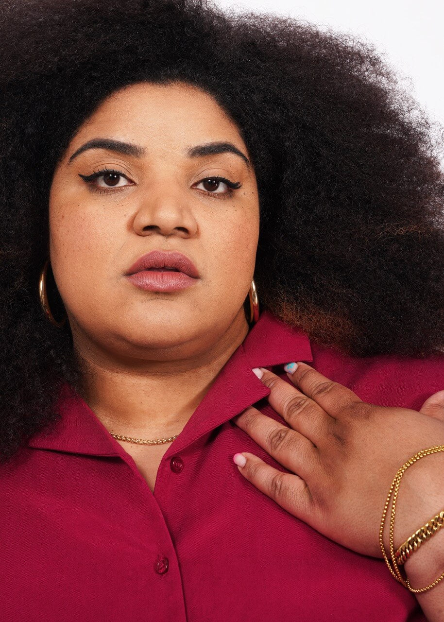 1X= A person with curly hair, wearing gold hoop earrings, a gold necklace, and The Everyday Camp Button Up in ultra-soft red fabric, confidently looks at the camera with one hand on their chest.