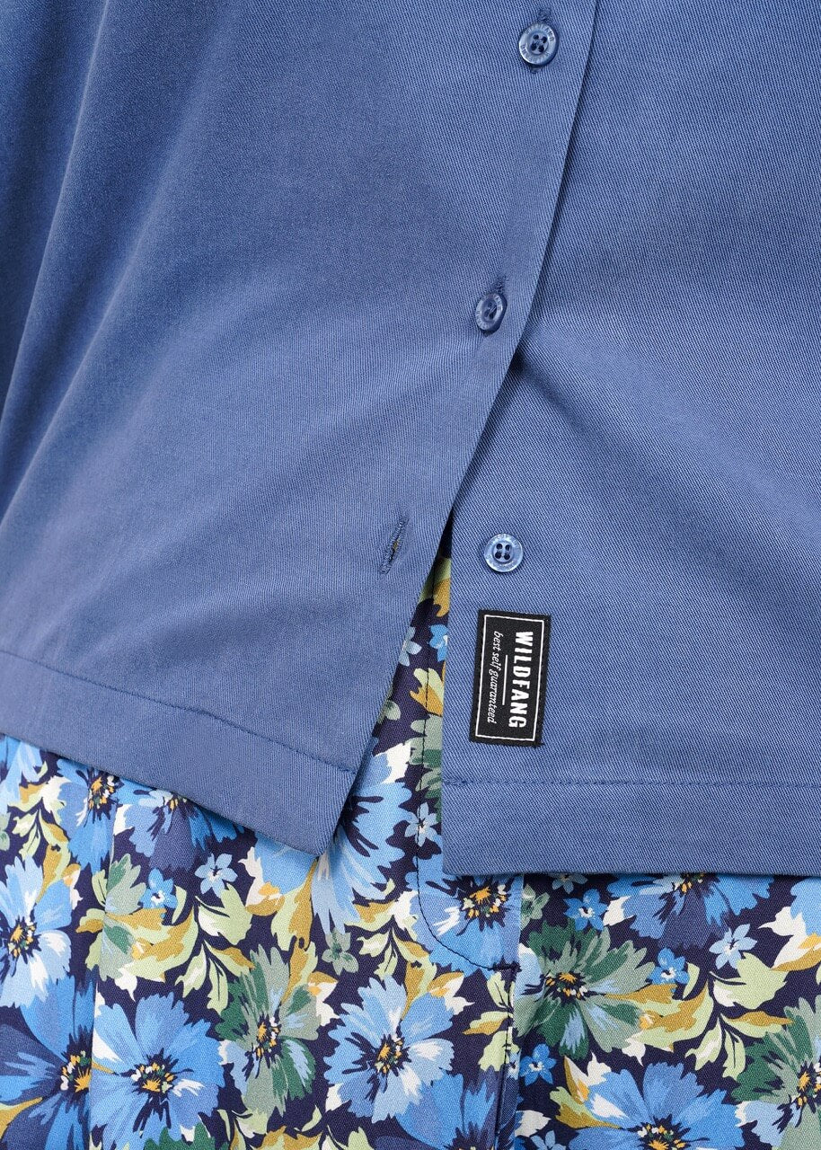 S=Close-up of The Everyday Camp Button Up, a blue camp collar shirt with buttons and visible label, paired with a skirt or pants in a vibrant blue, green, and yellow floral pattern. Relaxed fit crafted from soft cotton/Tencel fabric.