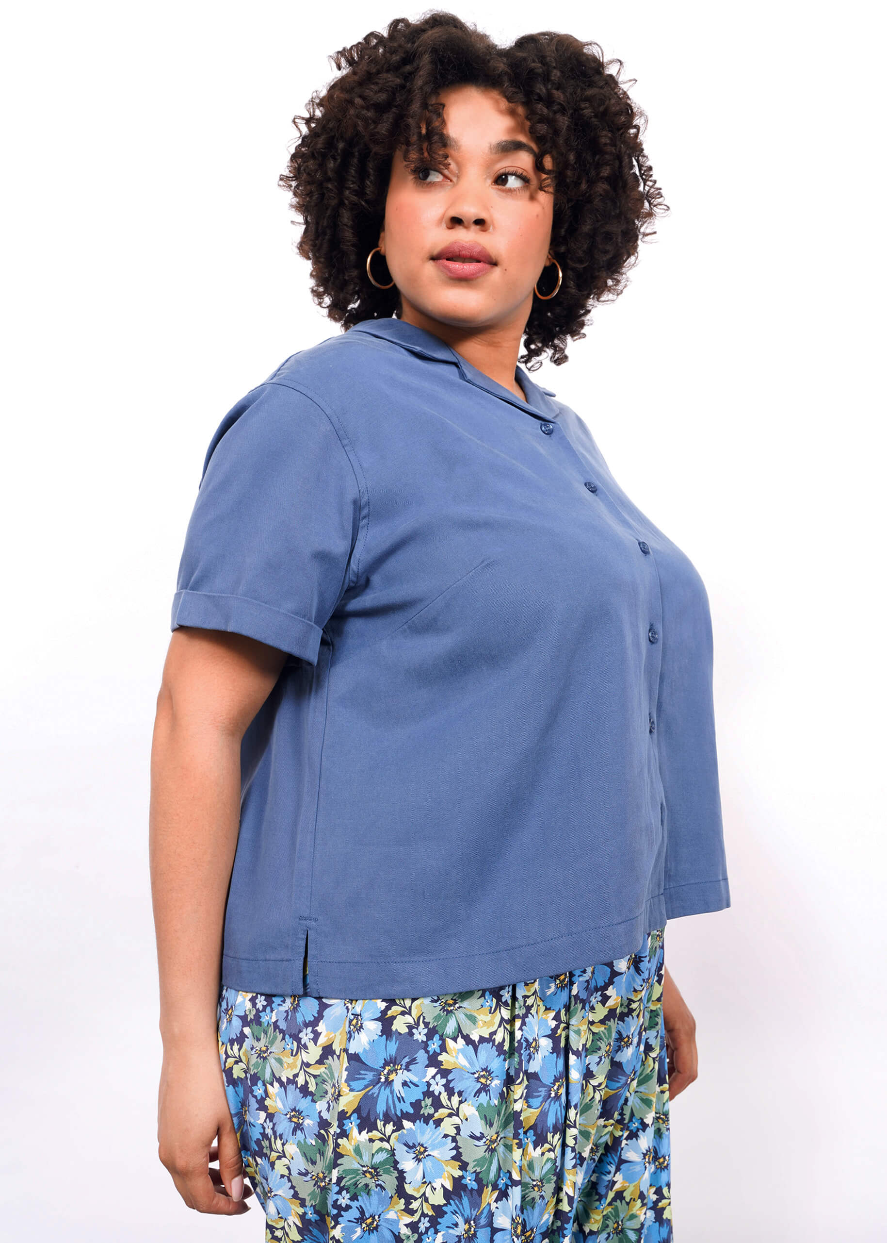 1X= A woman with curly hair looks to the side, wearing The Everyday Camp Button Up in blue and a floral skirt with blue, green, and yellow hues on a white background.