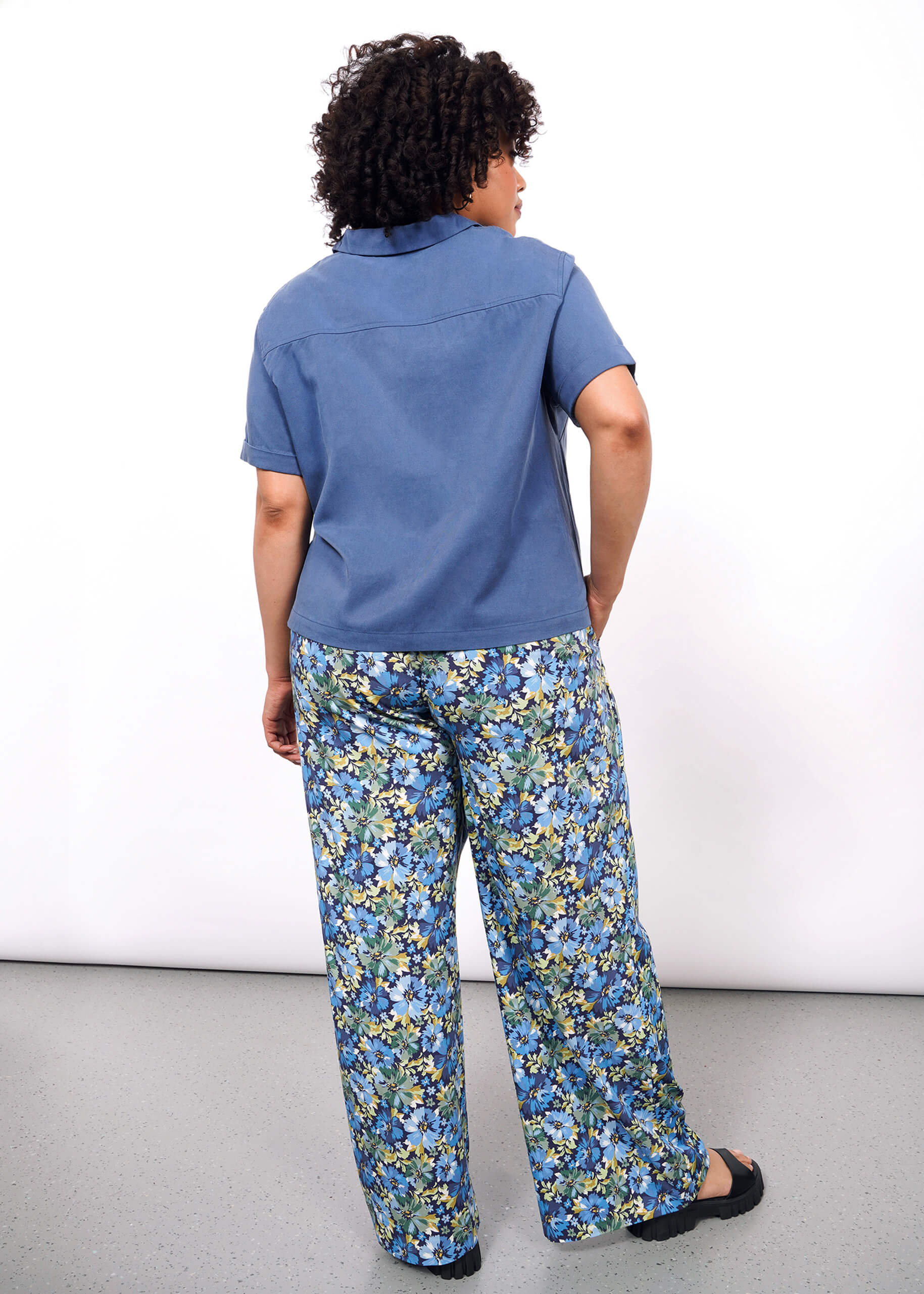 1X=A person with curly hair faces away, wearing The Everyday Camp Button Up in blue and loose, floral blue and green cotton/Tencel pants, set against a plain white background.