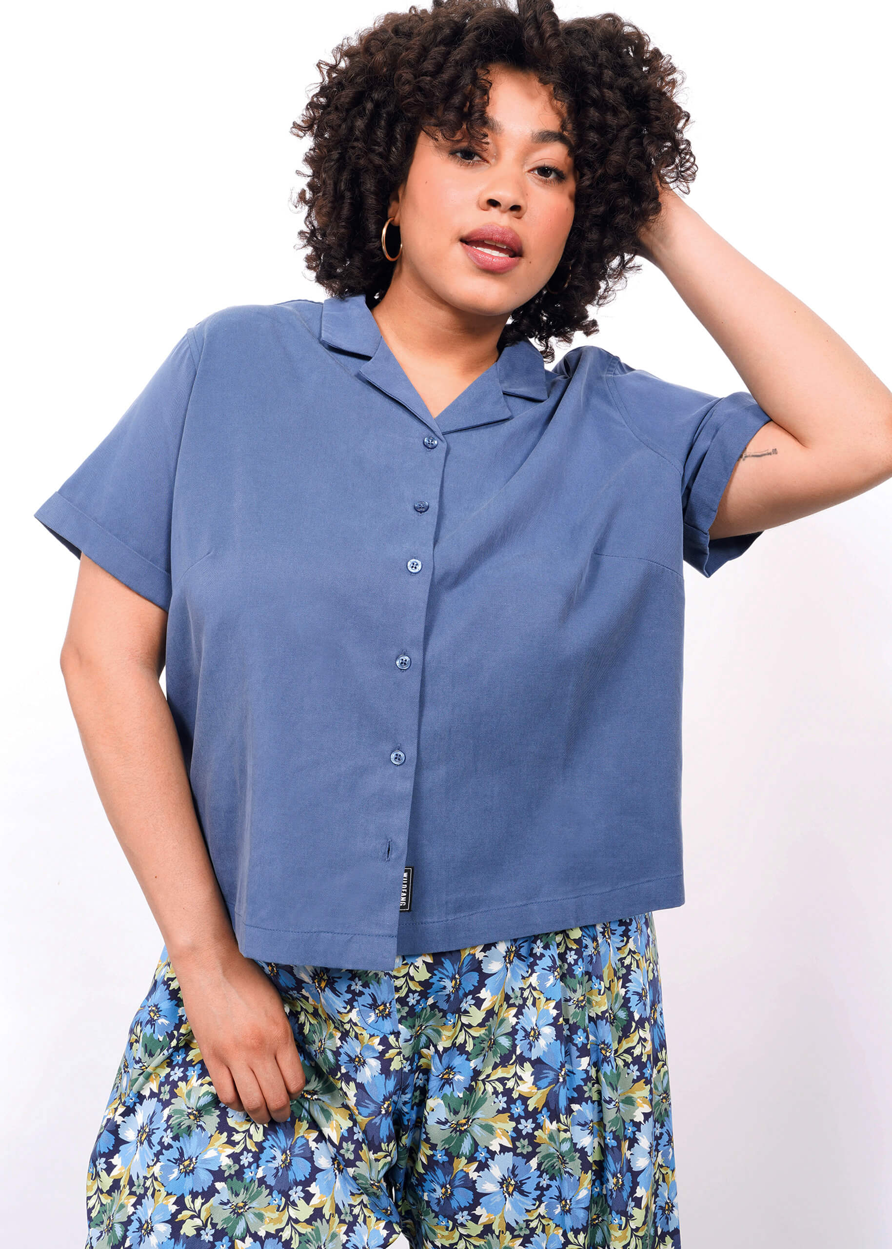 1X=A woman with curly hair wears The Everyday Camp Button Up, a relaxed fit blue shirt, paired with a floral skirt featuring blue, green, and yellow patterns, standing against a white background.