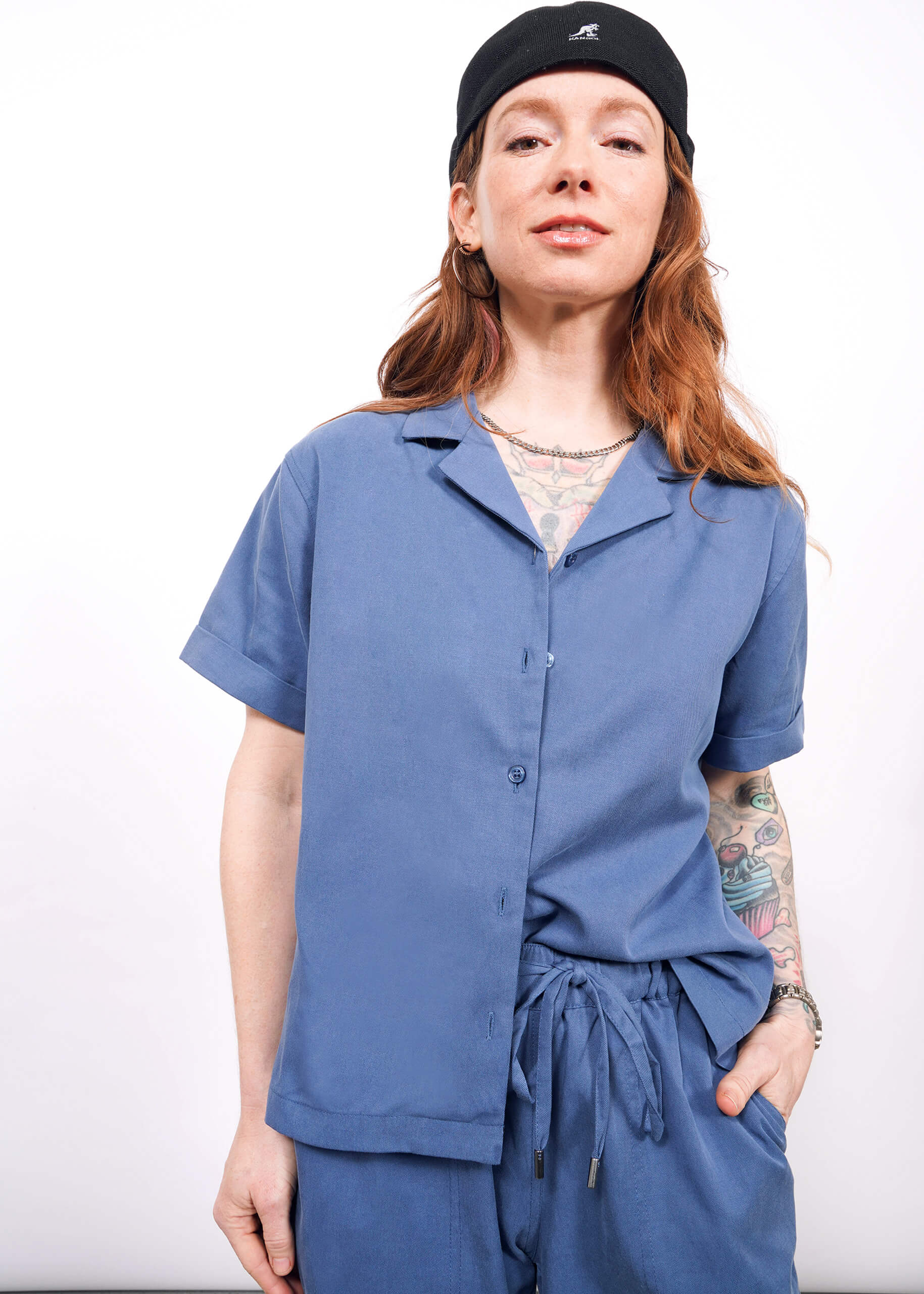 S=A person with long, wavy hair and visible tattoos wears a black cap and The Everyday Camp Button Up in blue with matching pants, standing against a white background with hand in pocket.