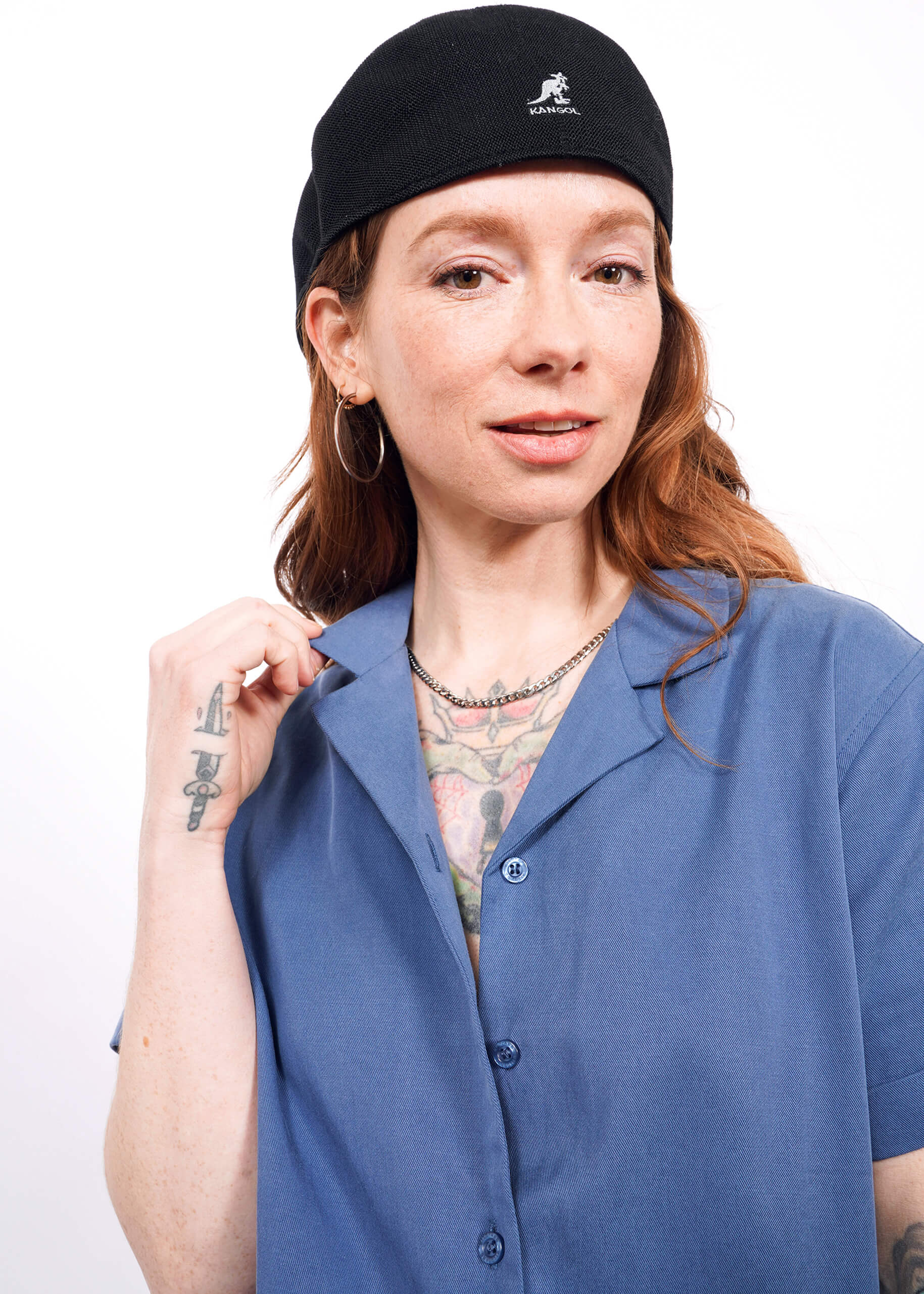 S= A woman with long reddish hair wears The Everyday Camp Button Up in blue with a black hat and hoop earrings. She has tattoos on her chest and hand, and smiles softly at the camera against a white background.