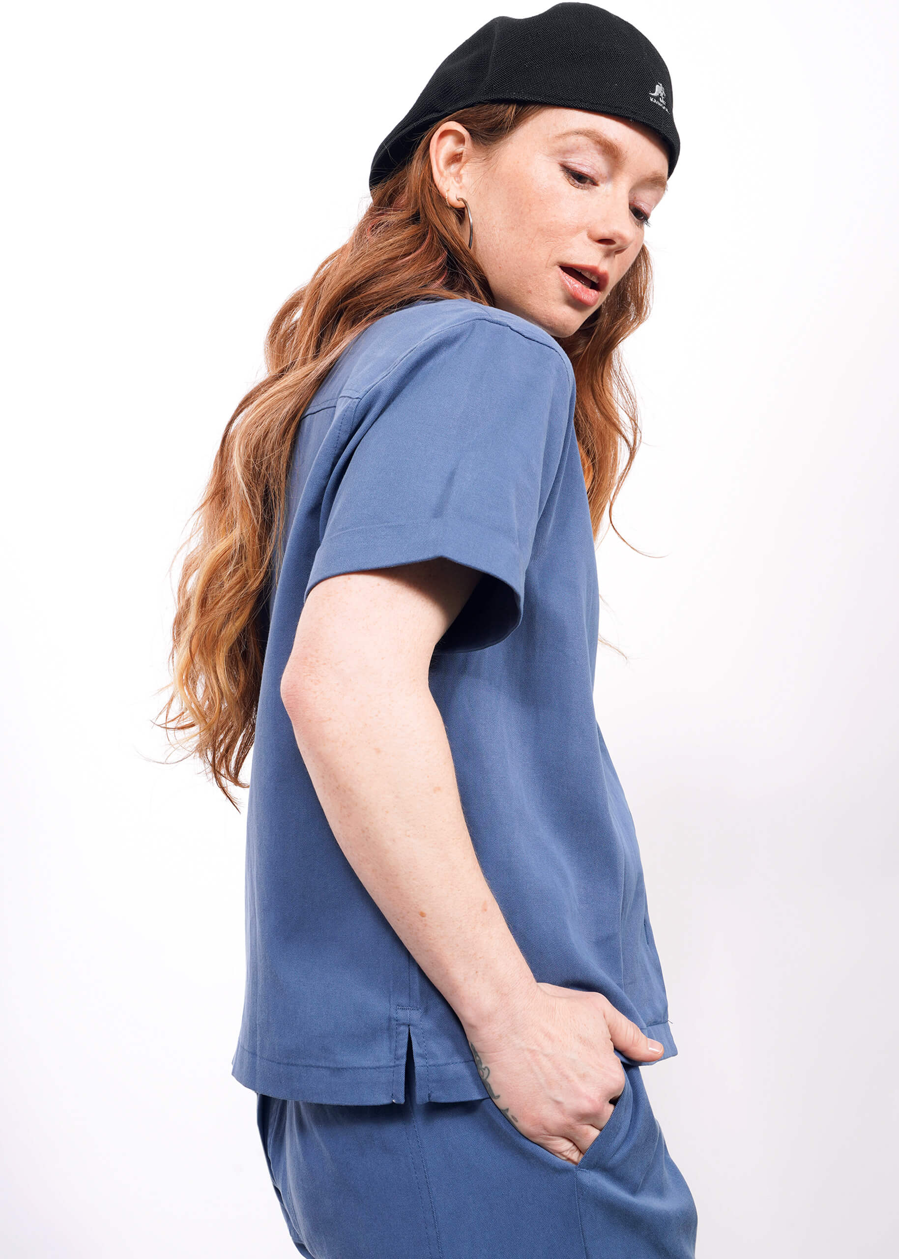S=Someone with long, wavy red hair wears The Everyday Camp Button Up in blue with matching pants and a black beret, standing against a white background and looking down to the side.