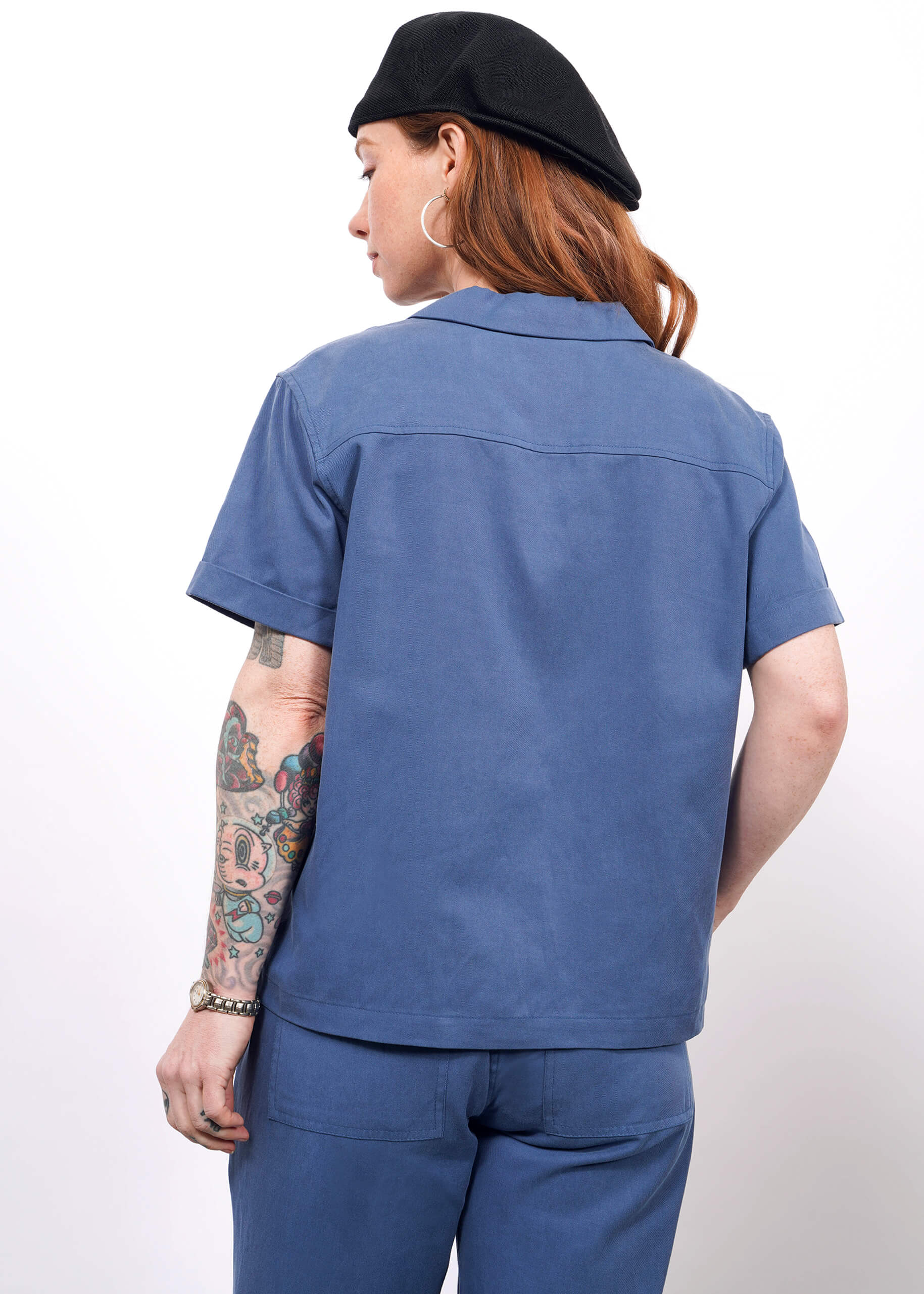 S= A person with long hair, a tattooed left arm, and facing away from the camera wears The Everyday Camp Button Up in relaxed blue with matching pants and a black beret, posed against a plain white background.