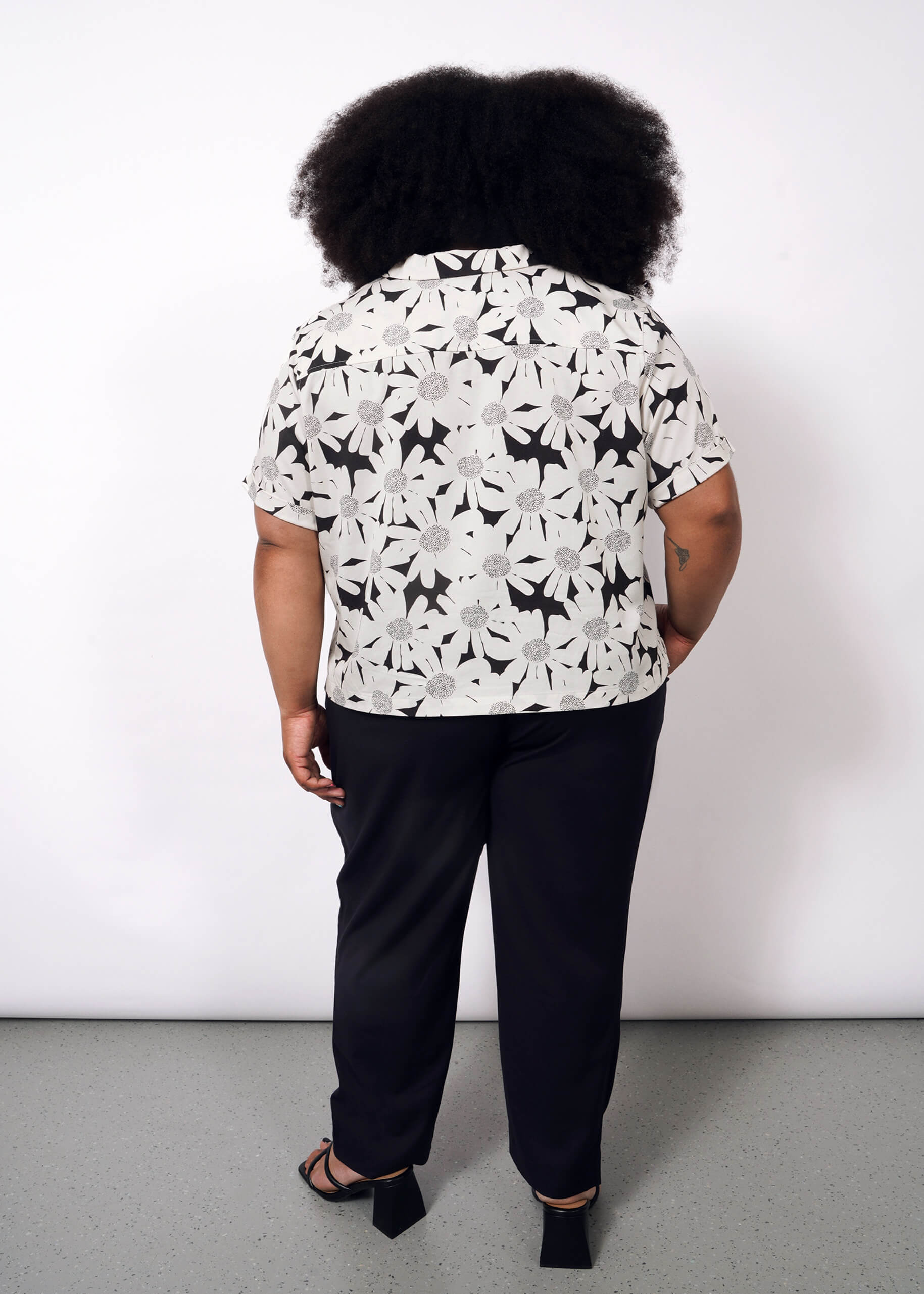 1X = A person with curly hair faces away, wearing The Everyday Camp Button Up—a relaxed-fit, black-and-white floral shirt in ultra-soft fabric—paired with black pants and sandals, against a plain light background.