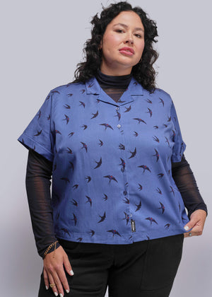 1X=Woman wearing a blue patterned shirt with black sleeves against a gray background