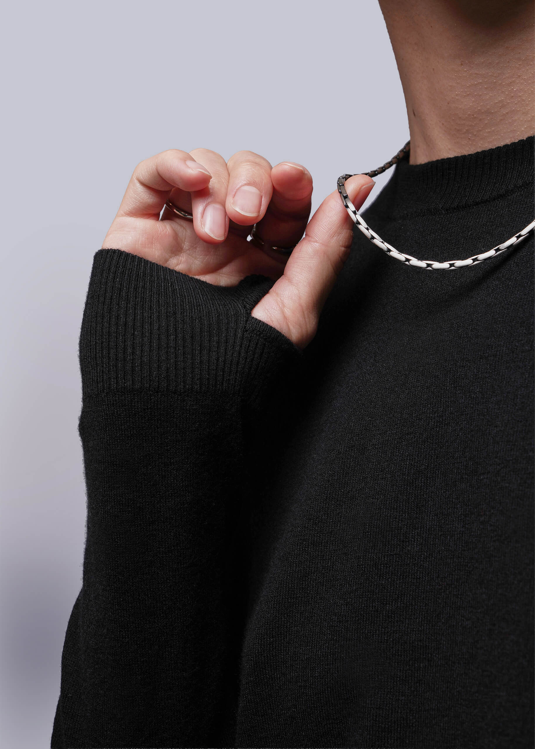 S=Person wearing a black sweater with a silver chain necklace against a light purple background