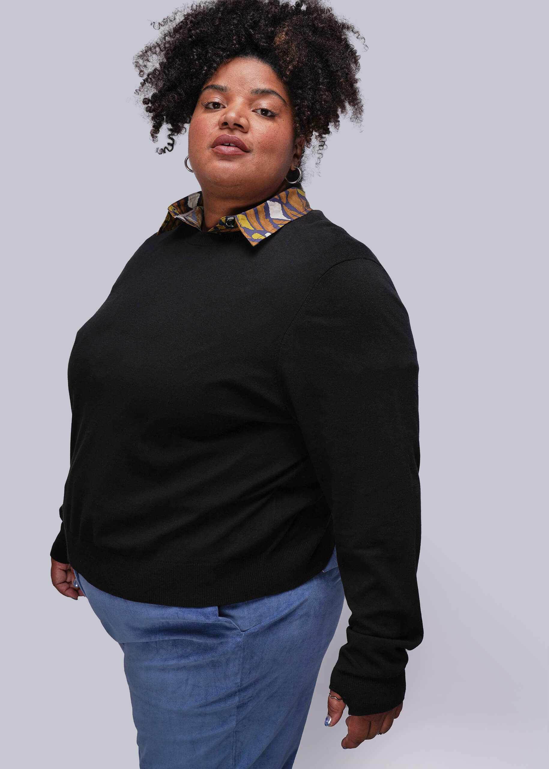 1X=Person wearing a black sweater with a colorful button up underneath against a white background