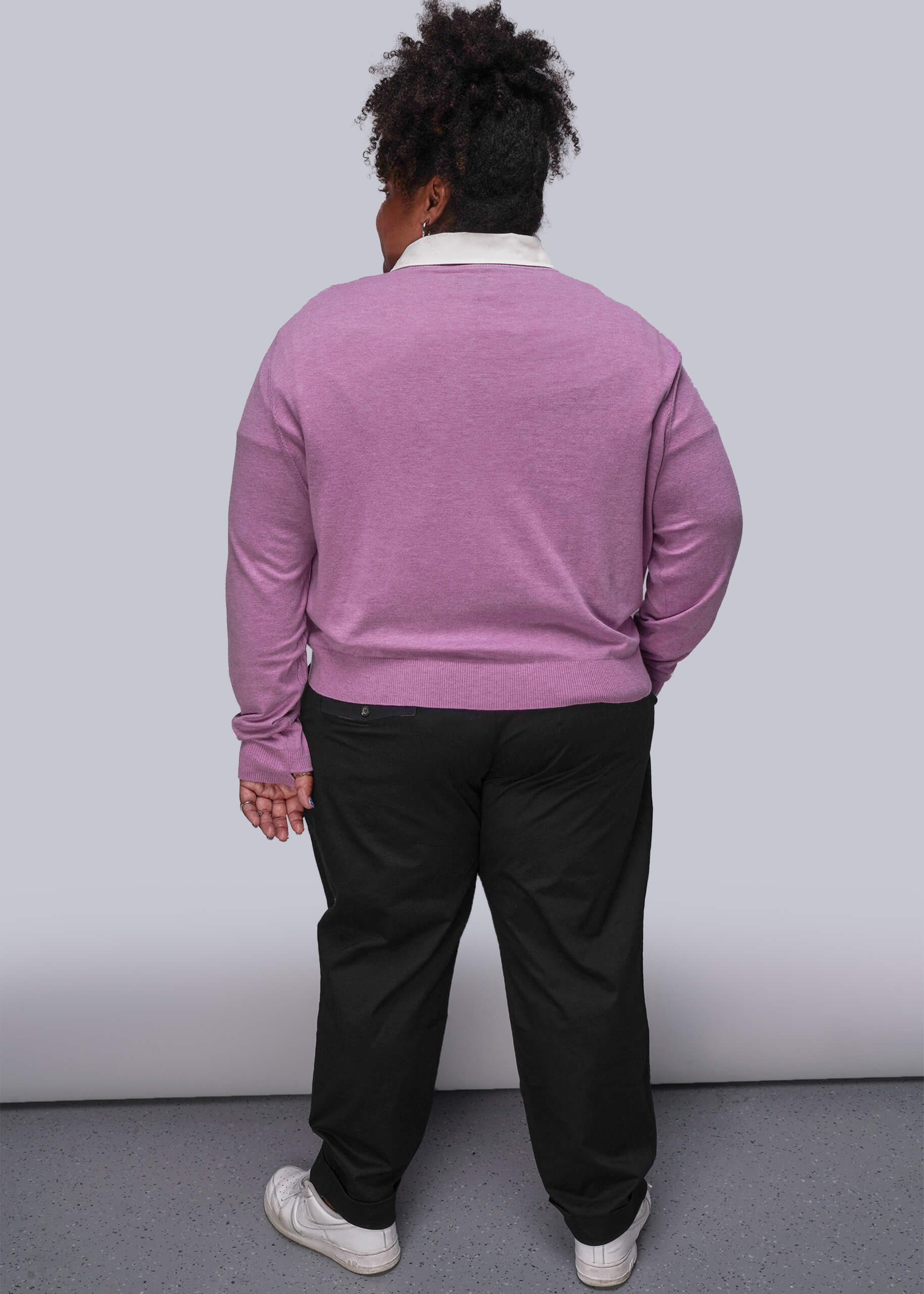 1X=Back view of person wearing a pink sweater and black pants against a white wall