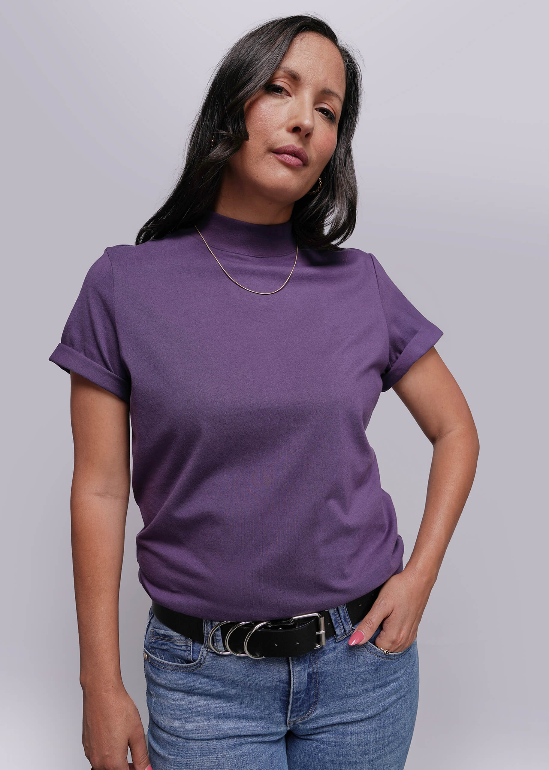 S=Woman wearing a purple turtleneck shirt and blue jeans against a plain background