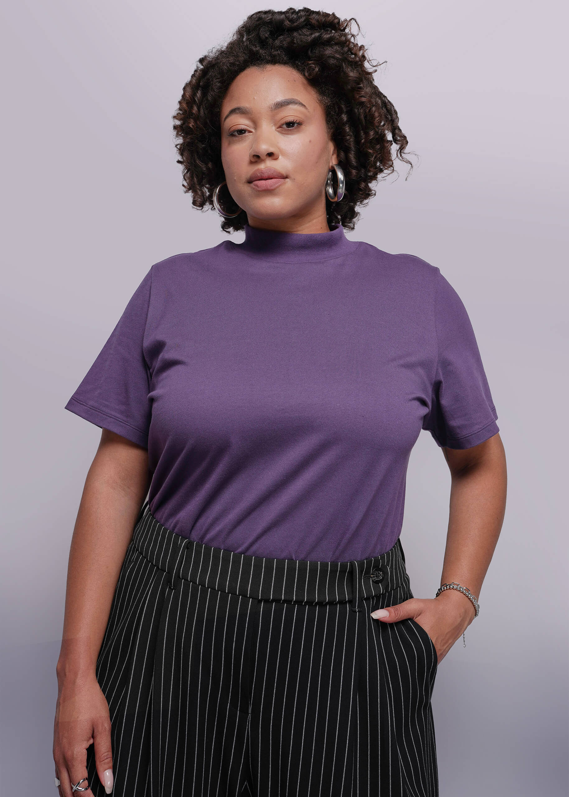 1X=Woman wearing a purple t-shirt and black pinstripe pants against a plain background