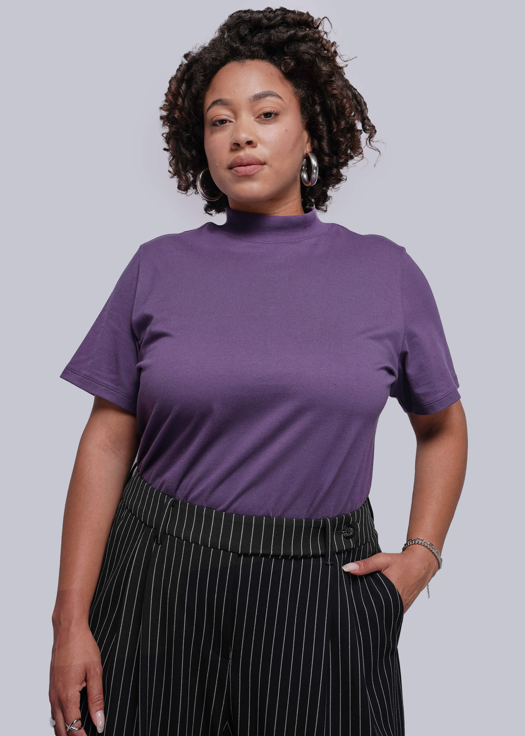 1X=Woman wearing a purple t-shirt and black pinstripe pants against a plain background