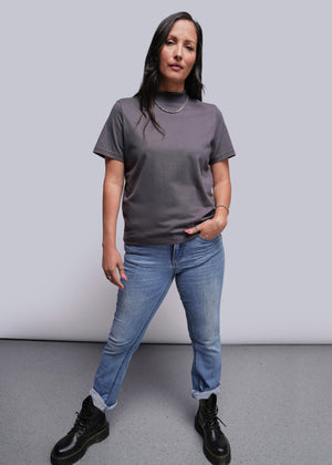 S=Person wearing a gray t-shirt, blue jeans, and black boots against a plain background