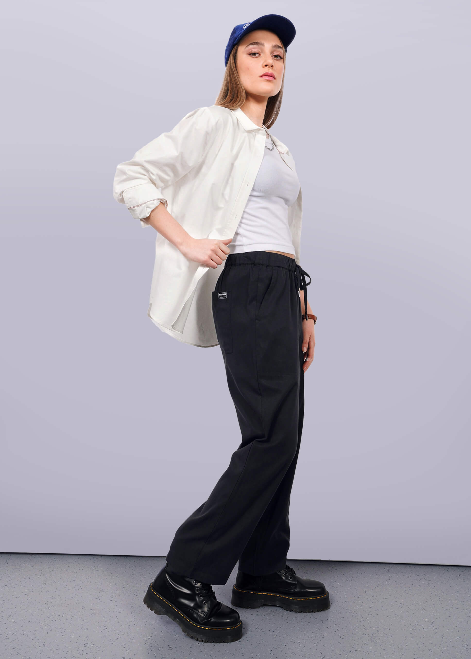 S=Wearing The Everyday Elastic Waist Pant in black, paired with a blue cap, white shirt over a white tank top, and black platform shoes, a person stands against a plain white background and looks confidently at the camera.