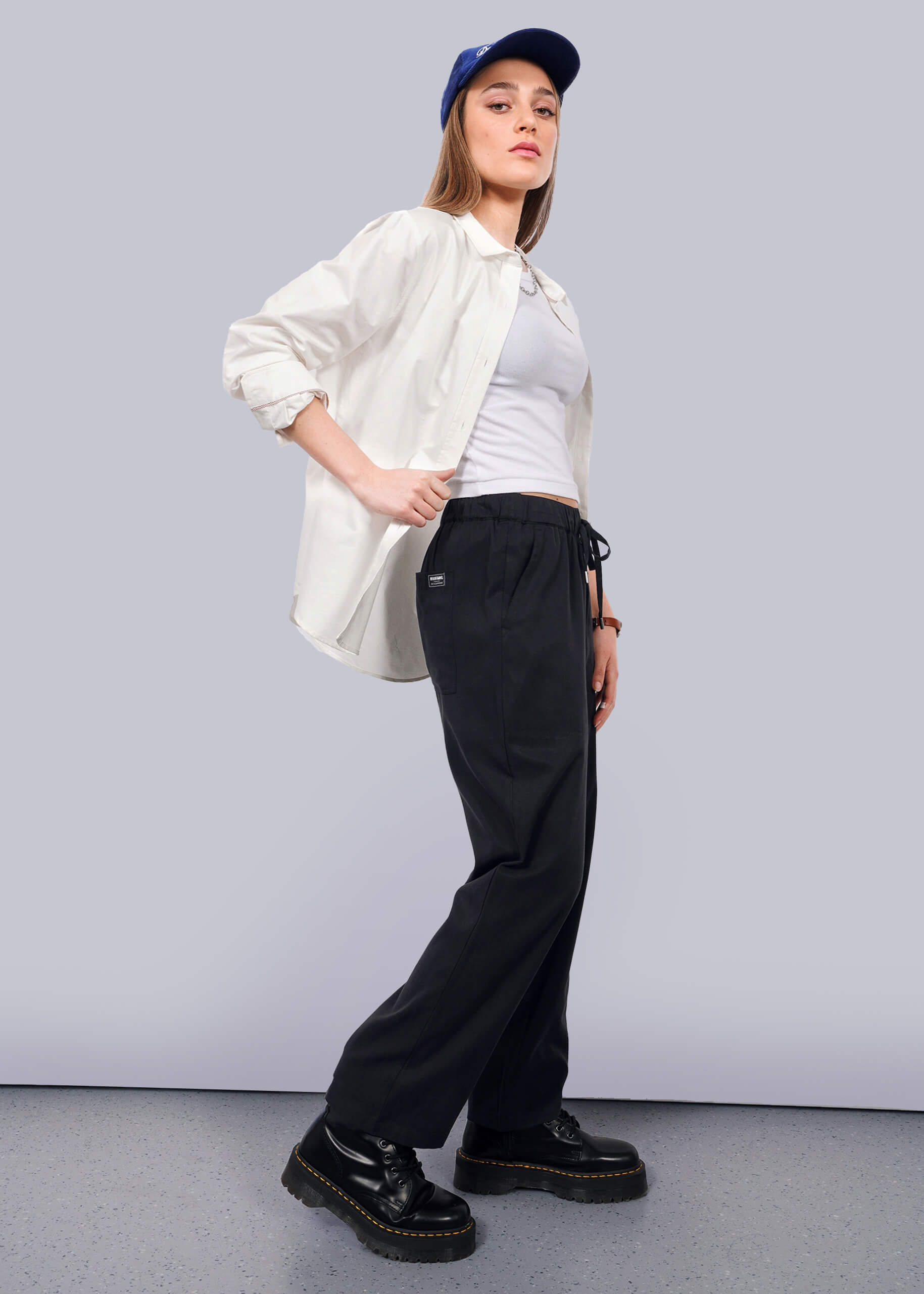S=Wearing The Everyday Elastic Waist Pant in black, paired with a blue cap, white shirt over a white tank top, and black platform shoes, a person stands against a plain white background and looks confidently at the camera.