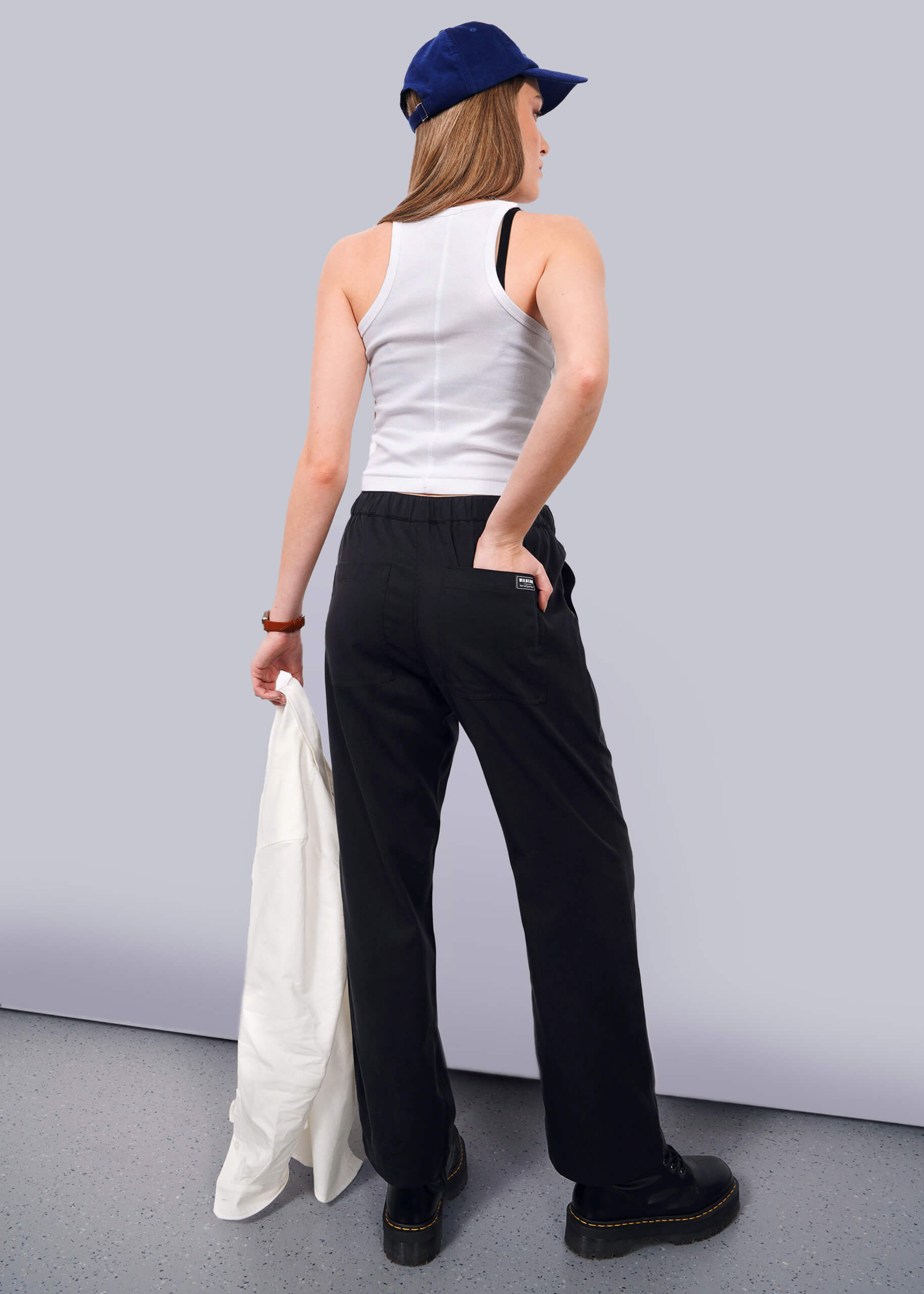 S=A person stands facing away against a white background, wearing The Everyday Elastic Waist Pant in black, paired with a blue cap, white tank top, black shoes, and holding a white jacket.