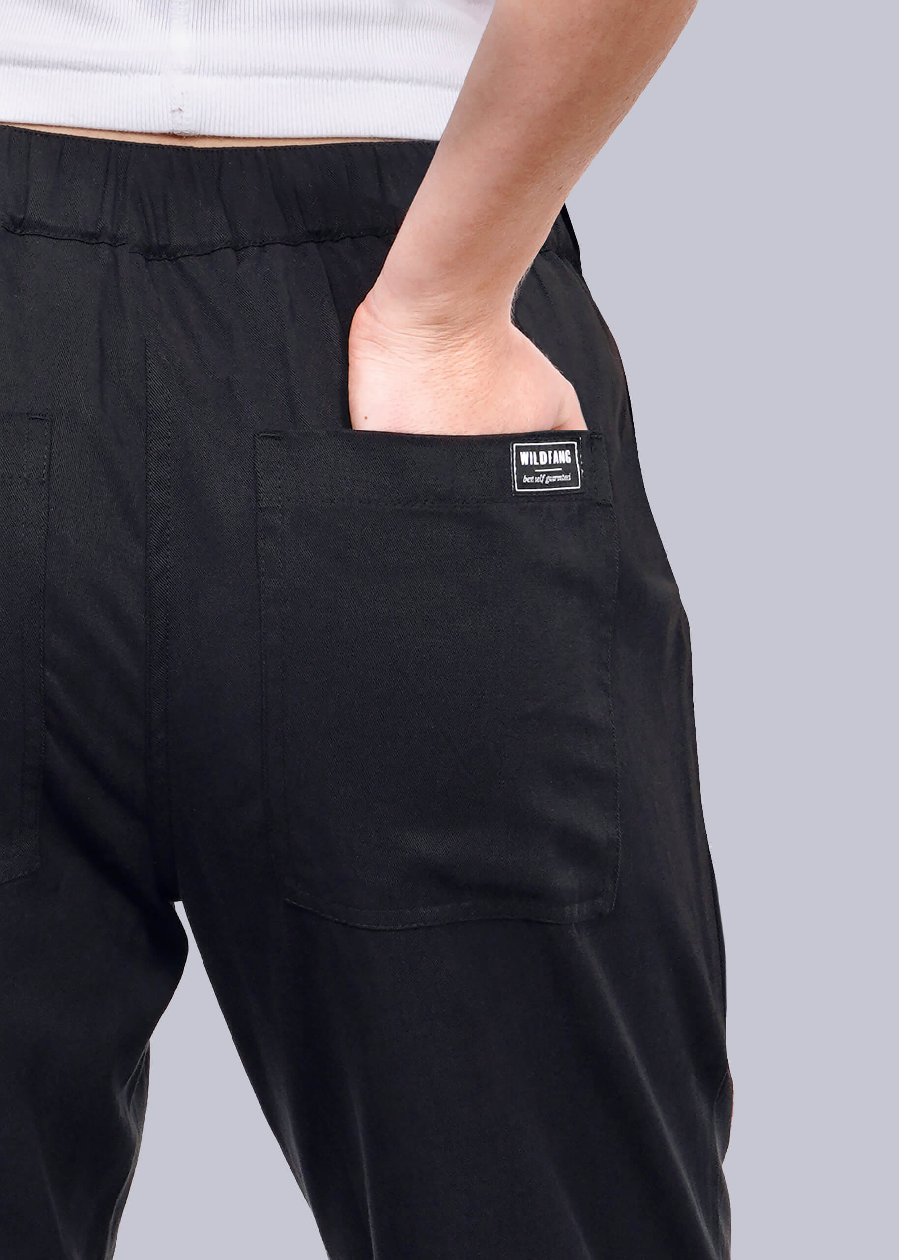 S= A person wears The Everyday Elastic Waist Pant in black with a white shirt, their right hand in the back pocket. A small black label on the pocket reads WILD BANG in white. The background is plain white.