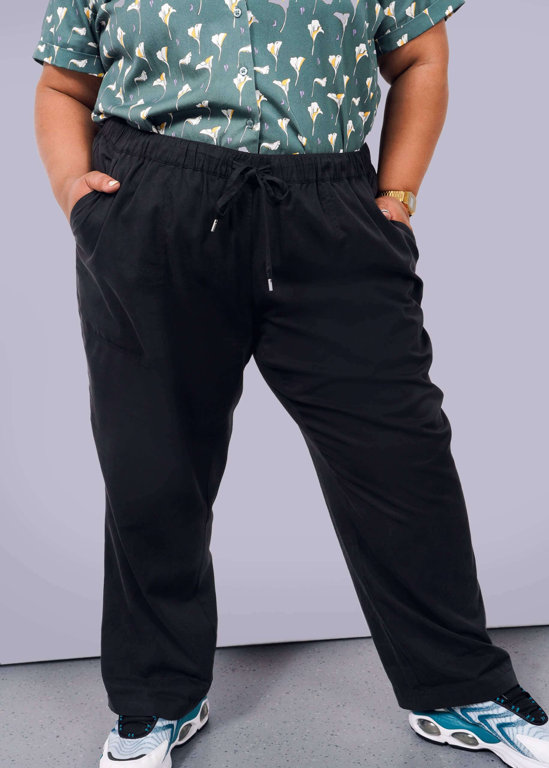 1X= A person stands with hands in their pockets against a plain white background, wearing a green patterned shirt, The Everyday Elastic Waist Pant in black, white and blue sneakers, and a gold watch.