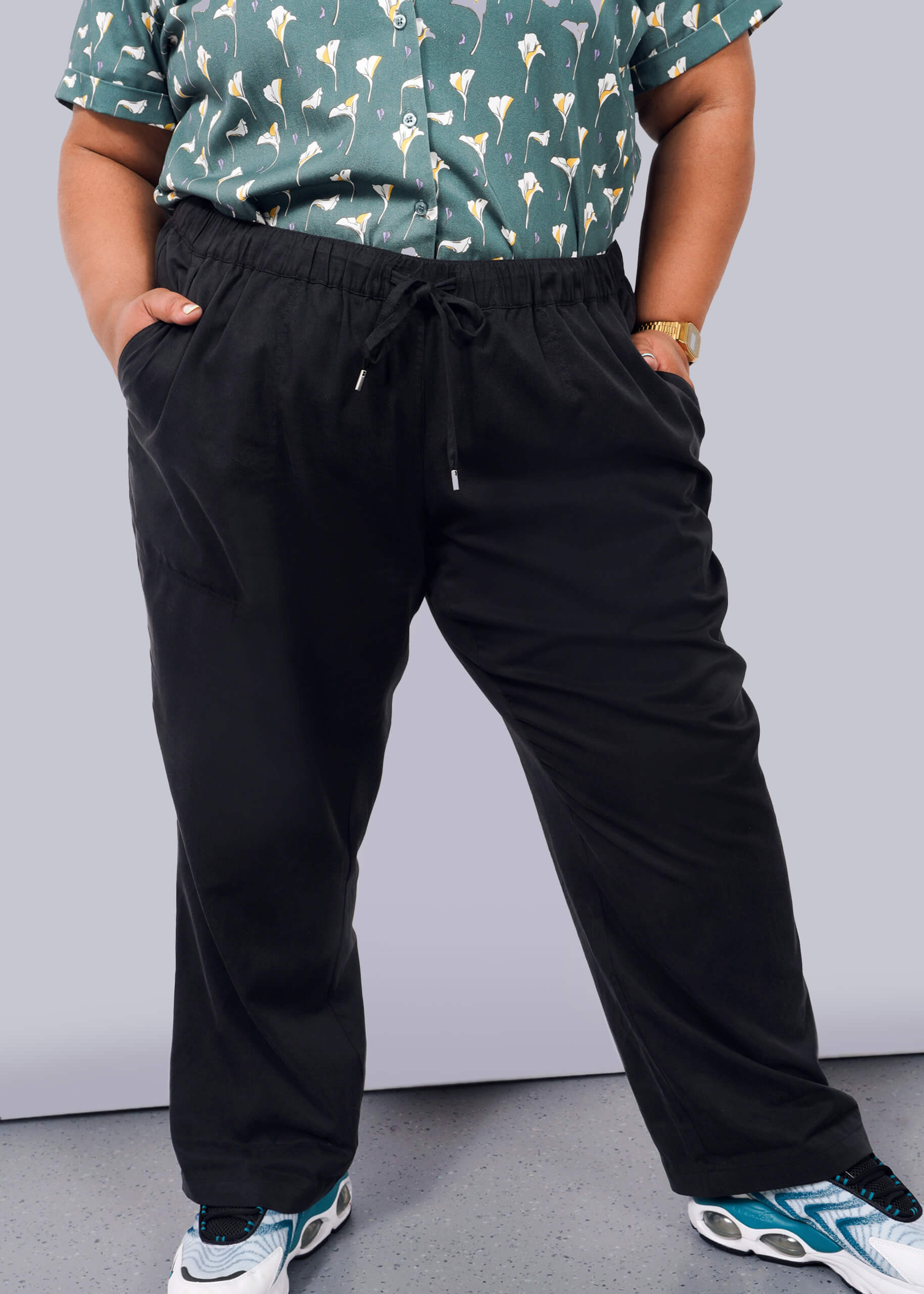 1X= A person stands with hands in their pockets against a plain white background, wearing a green patterned shirt, The Everyday Elastic Waist Pant in black, white and blue sneakers, and a gold watch.