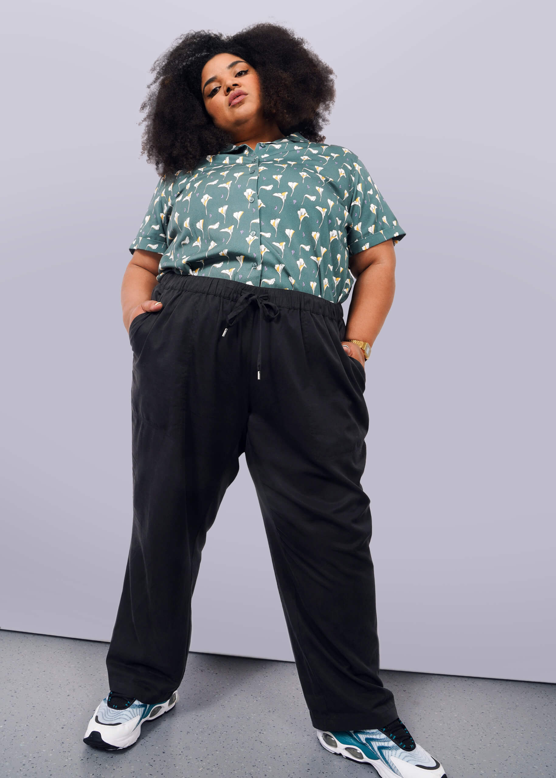 1X=A person with voluminous curly hair stands confidently against a white background, wearing a green patterned short-sleeve shirt, The Everyday Elastic Waist Pant in black, a watch, and white, black, and teal sneakers.