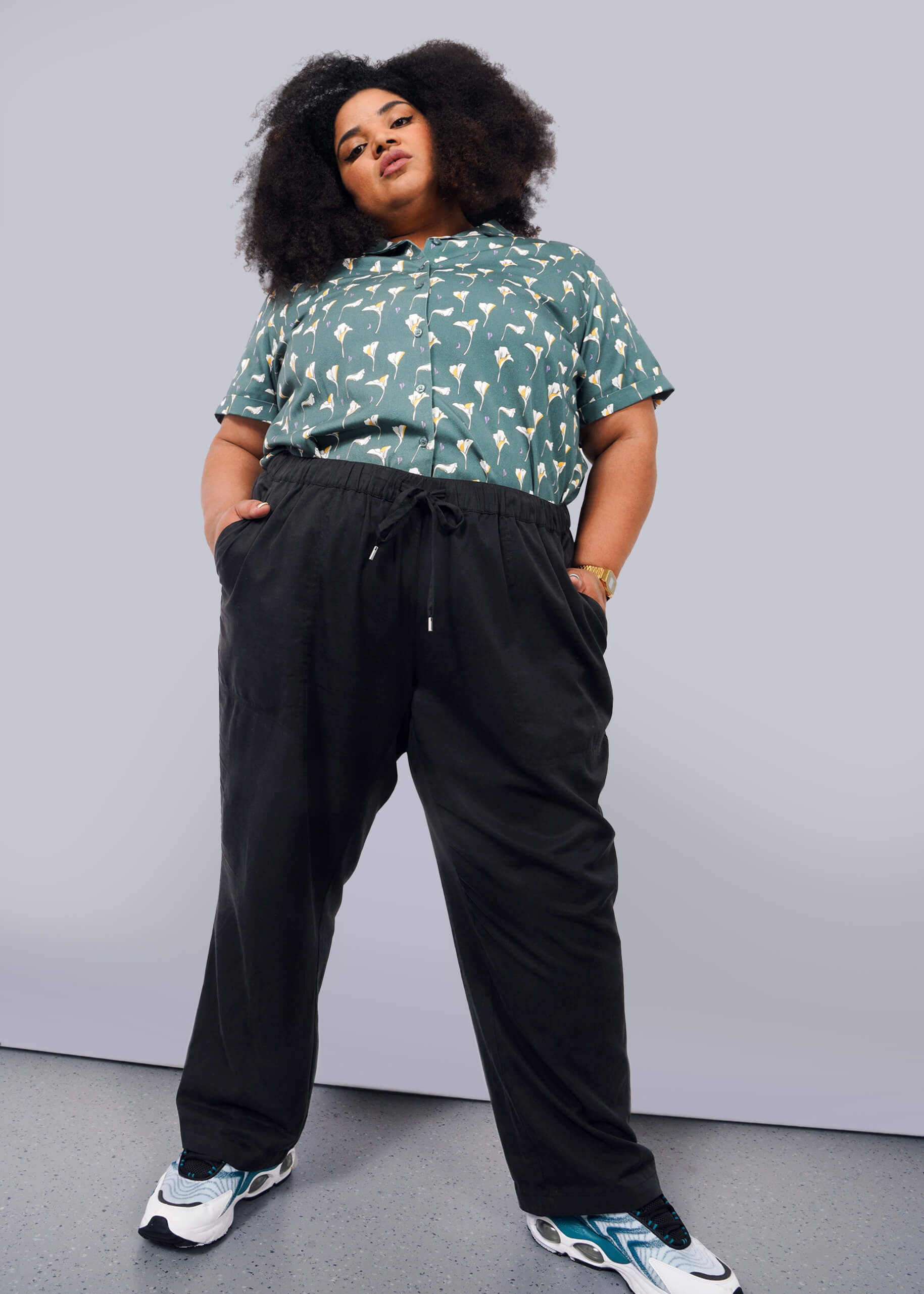 1X=A person with voluminous curly hair stands confidently against a white background, wearing a green patterned short-sleeve shirt, The Everyday Elastic Waist Pant in black, a watch, and white, black, and teal sneakers.