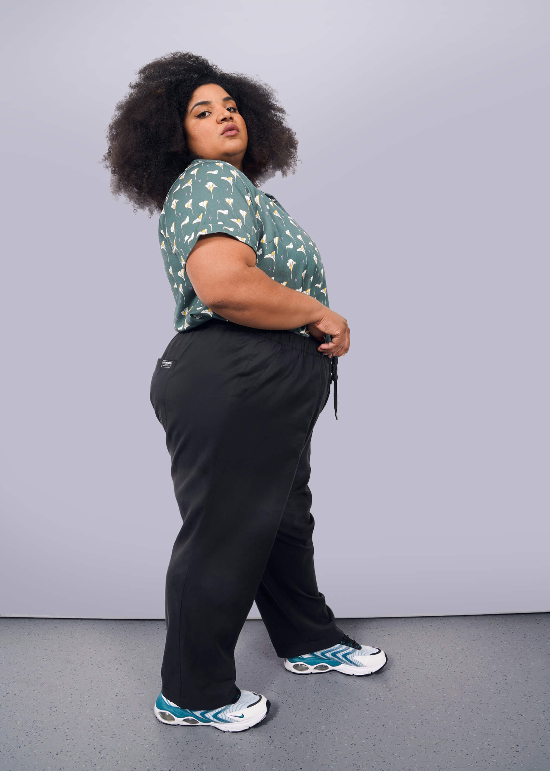 1X= A person with curly hair stands sideways against a white background, wearing a green patterned shirt, The Everyday Elastic Waist Pant in black, and white sneakers with blue accents.
