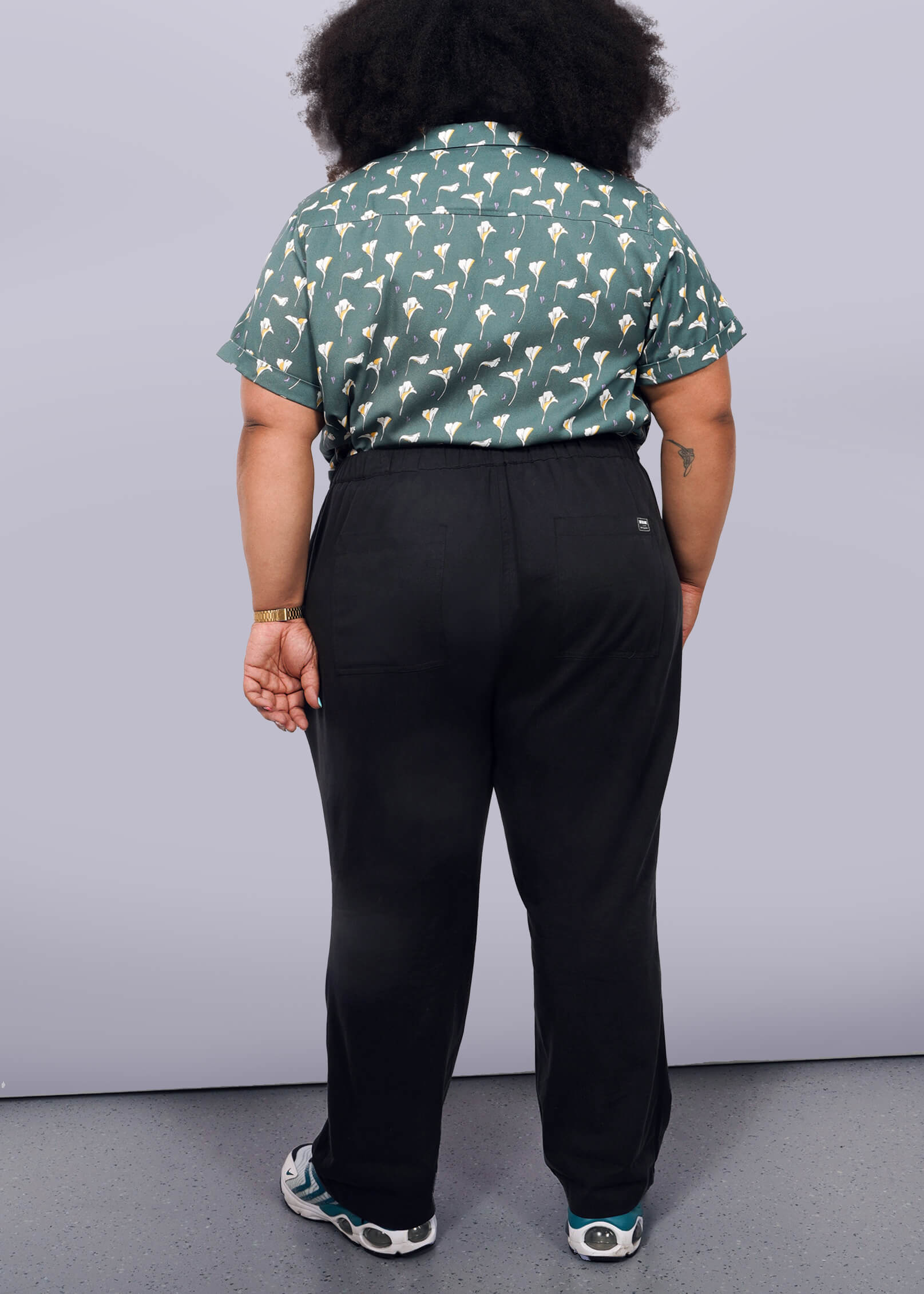 1X=A person with curly hair faces away from the camera, wearing a green patterned short-sleeve shirt, The Everyday Elastic Waist Pant in black, and sneakers, set against a plain white background.