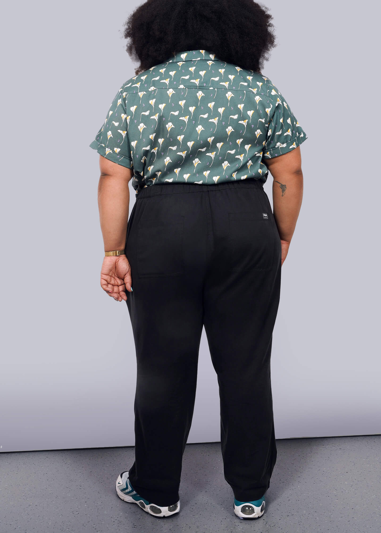 1X=A person with curly hair faces away from the camera, wearing a green patterned short-sleeve shirt, The Everyday Elastic Waist Pant in black, and sneakers, set against a plain white background.