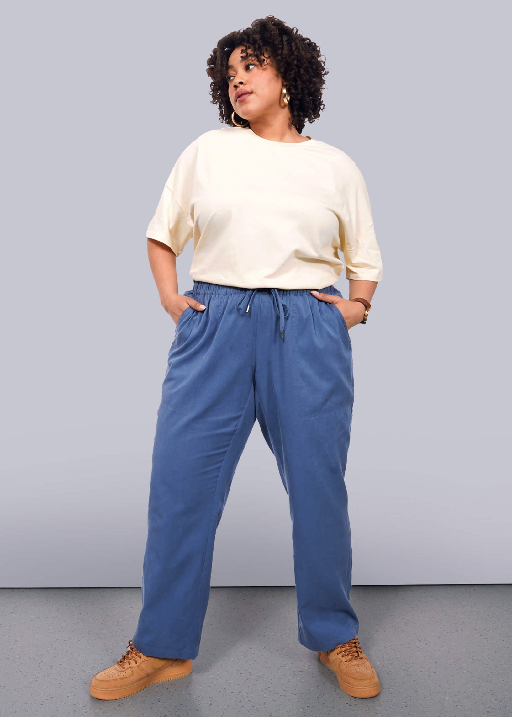 1X= A woman with curly hair stands against a white background, wearing The Everyday Elastic Waist Pant in blue with an elastic waistband and drawstring, paired with a loose cream top. She has her hands in her pockets and looks confidently to the side.