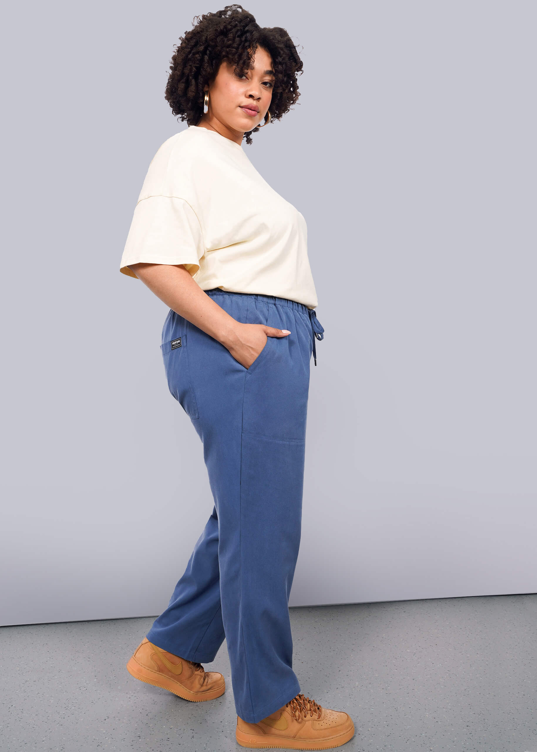 1X=A woman with curly hair stands in profile against a white background, wearing blue The Everyday Elastic Waist Pant with a relaxed fit and elastic waist, a loose cream T-shirt, tan sneakers, and her hands in her pockets.