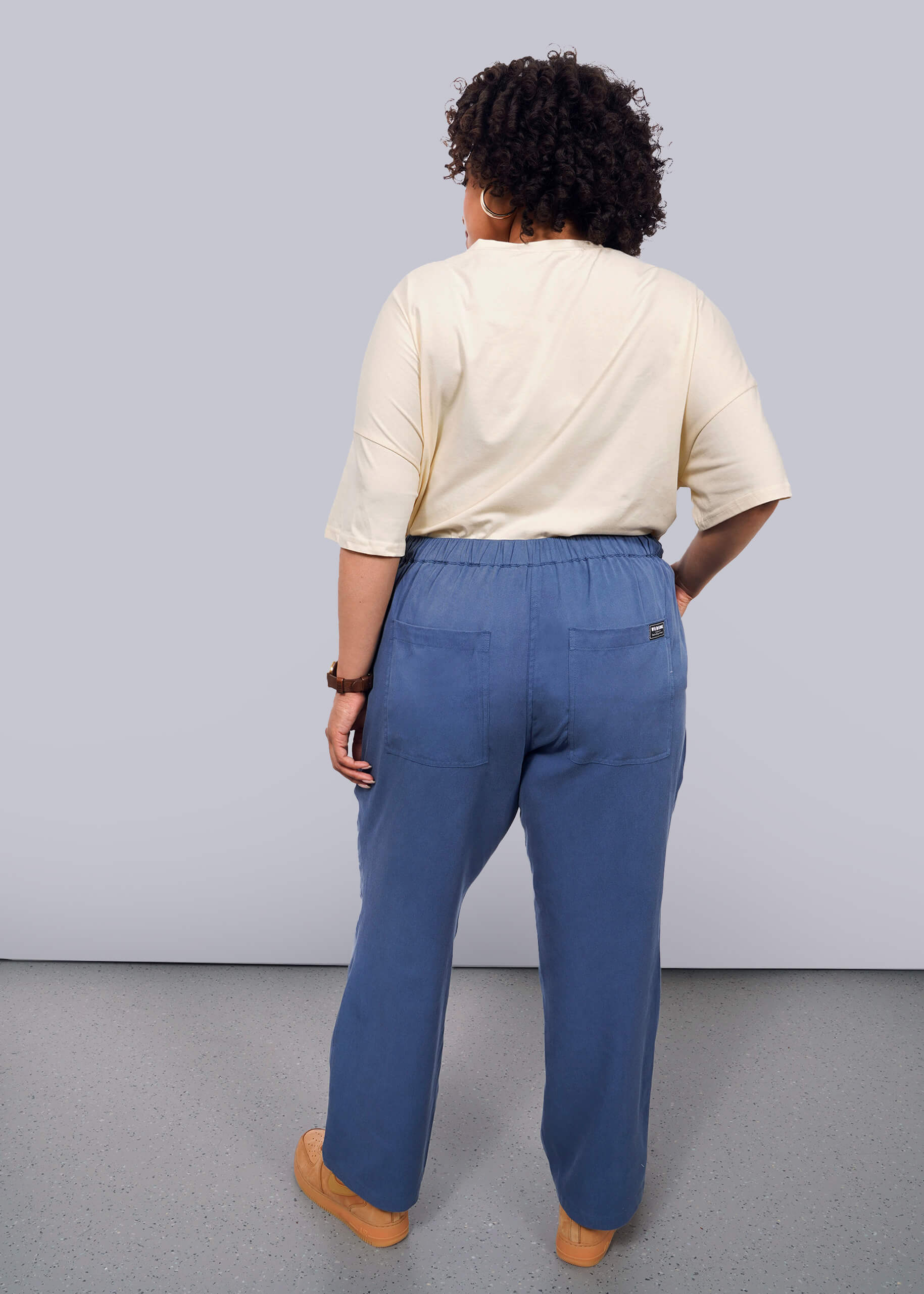 1X= A person with short curly hair, wearing a cream top and The Everyday Elastic Waist Pant in blue, stands facing a white wall with their back to the camera. They wear brown shoes and a bracelet.