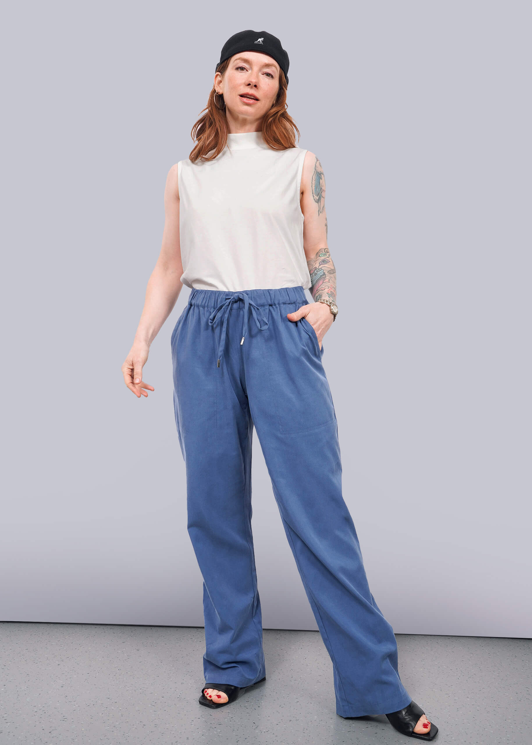 S=A woman wears The Everyday Elastic Waist Pant in blue with a white sleeveless top, black sandals, and a black cap. She stands against a white background, showing tattoos on her left arm and poses confidently with one hand in her pocket.