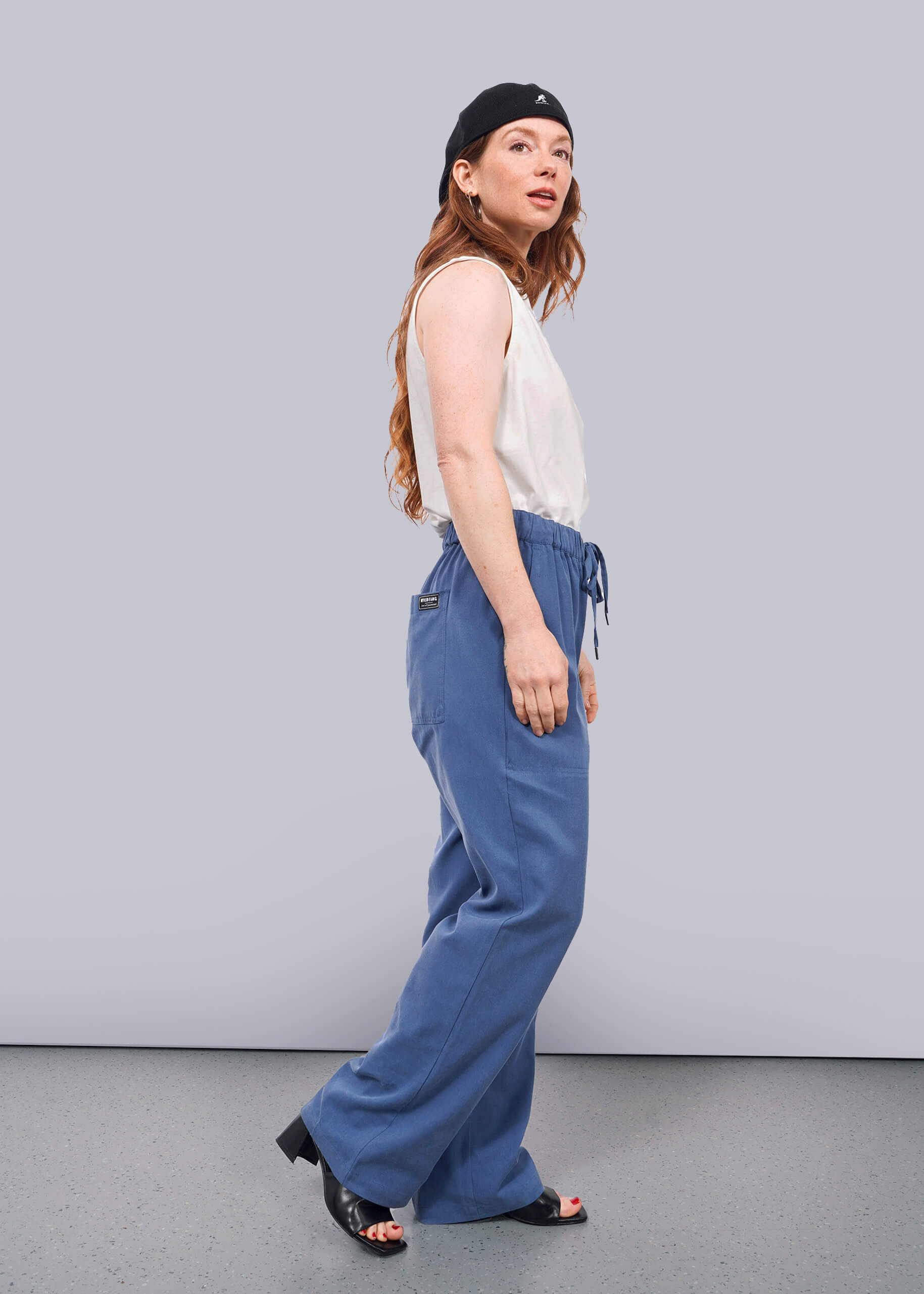 S=A woman with long hair wears The Everyday Elastic Waist Pant in blue, paired with a black beanie and a white sleeveless top, standing sideways and looking at the camera against a plain white background.