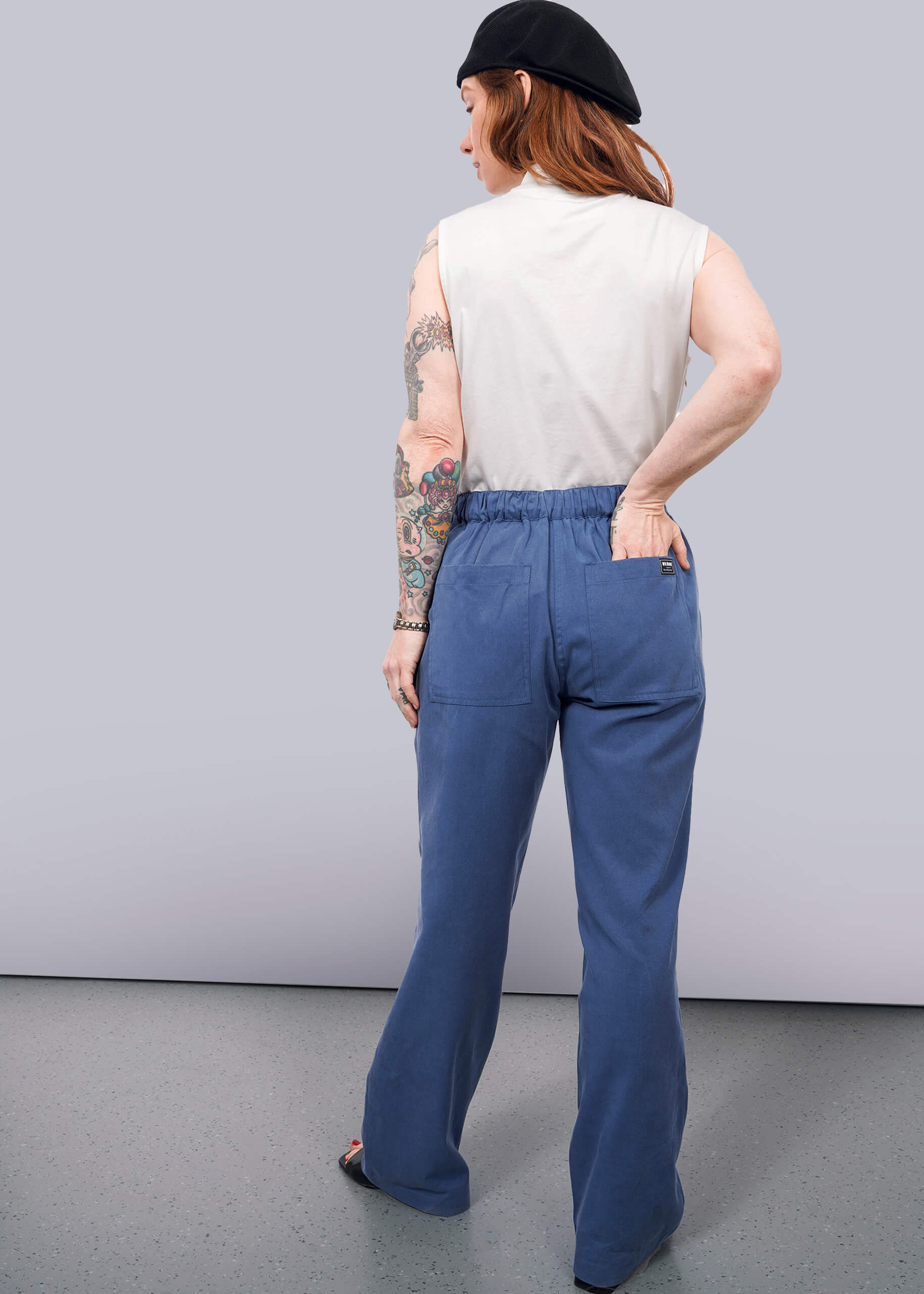 S= A person stands with their back to the camera, showing tattooed arms, wearing The Everyday Elastic Waist Pant in blue with a sleeveless white top and black beret against a plain light background.