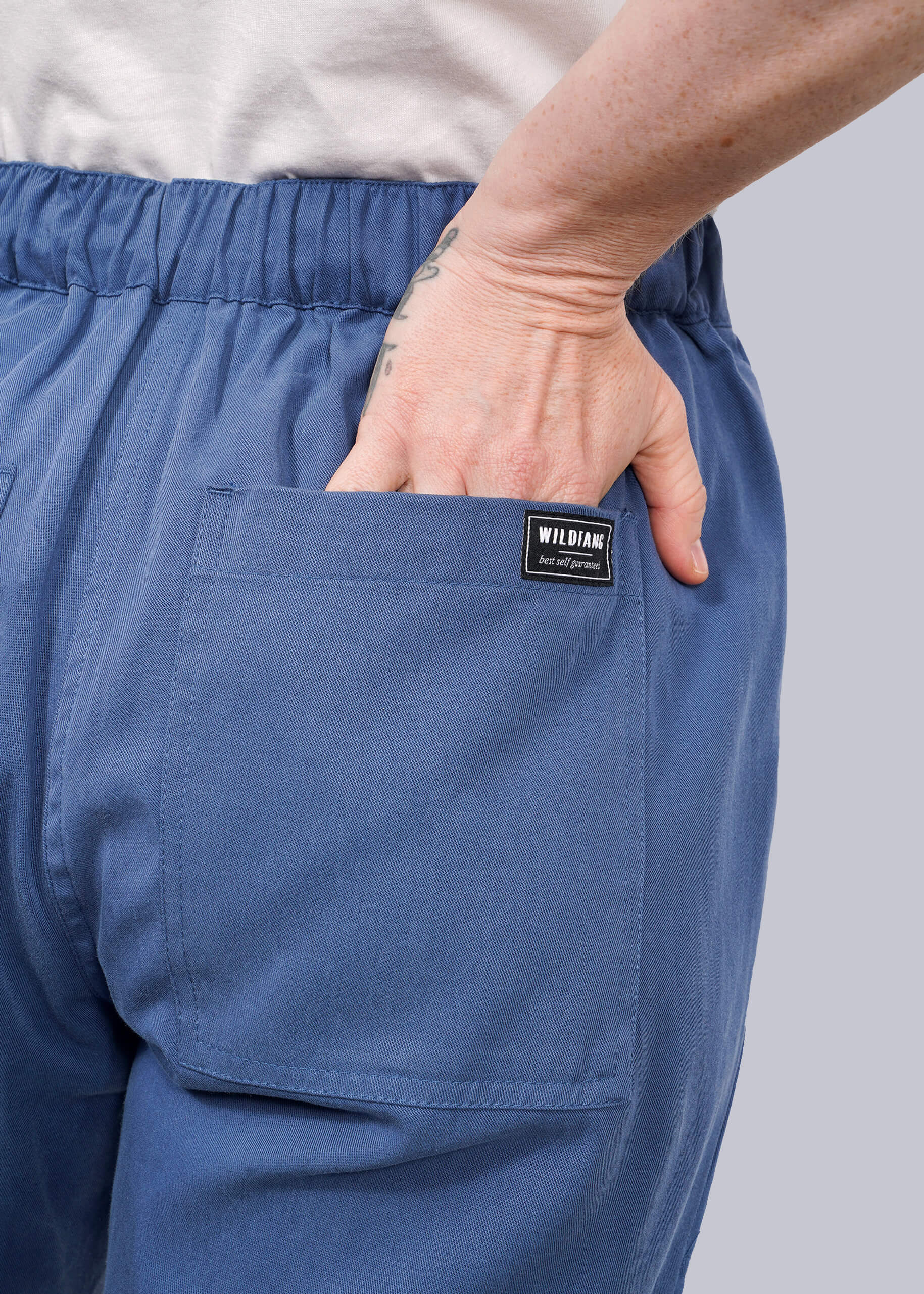 S=A person is shown from the back wearing The Everyday Elastic Waist Pant in blue, hand in the pocket with a visible WILDANG label, showing breathable fabric and a wrist tattoo.