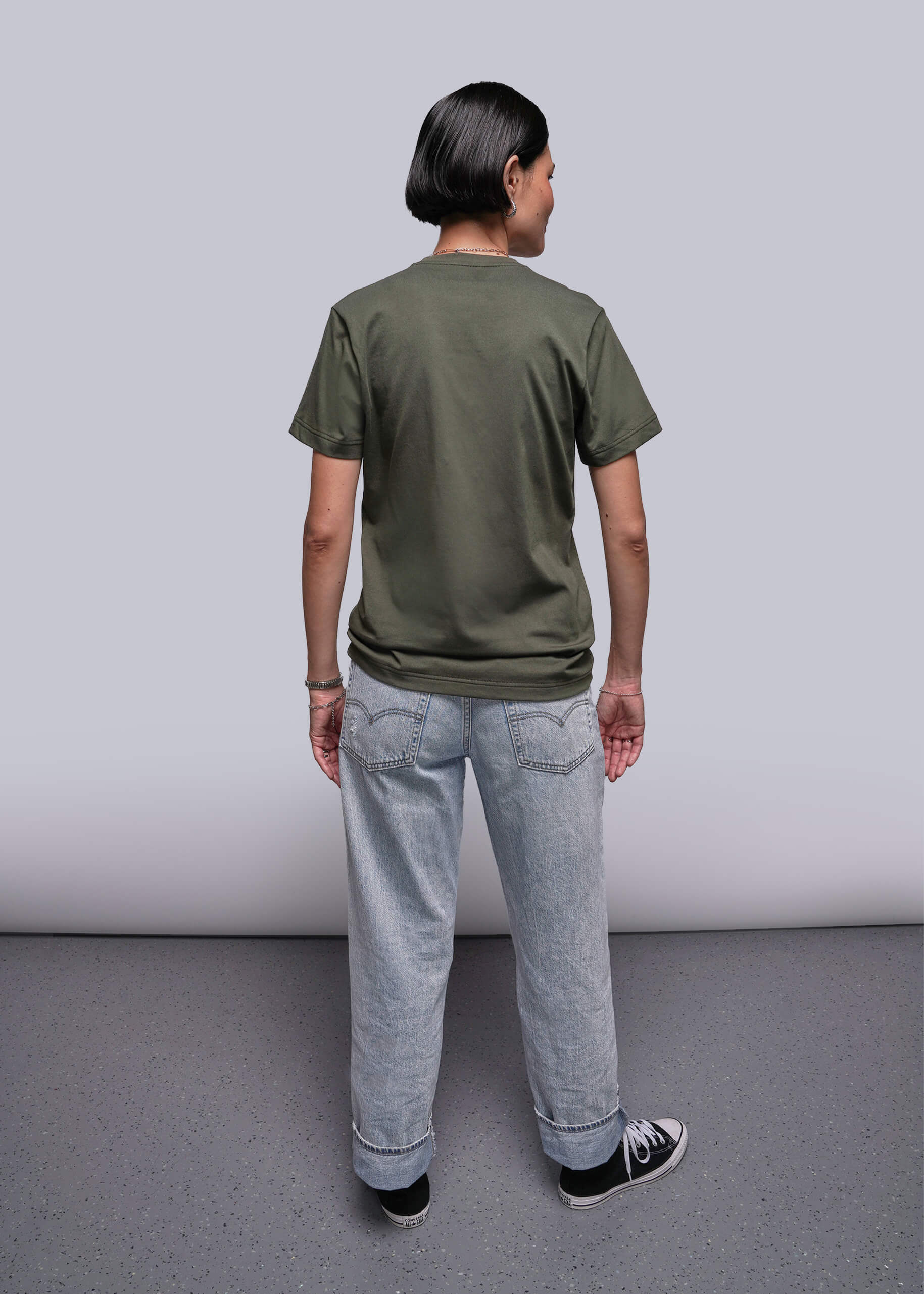 S=Back of person wearing an olive green t-shirt and light blue jeans standing against a plain background