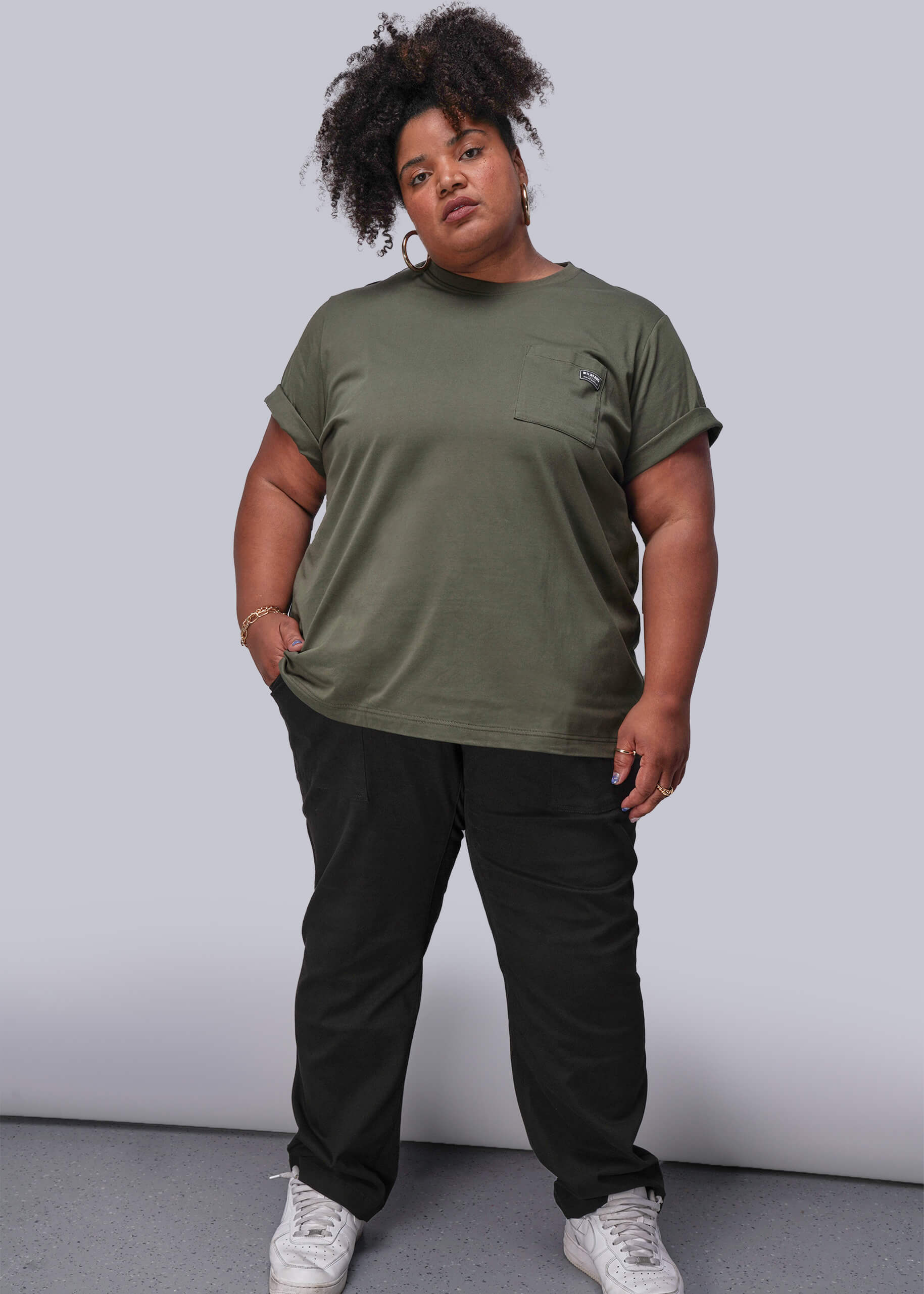 1X=Person wearing an olive green t-shirt and black pants against a plain background