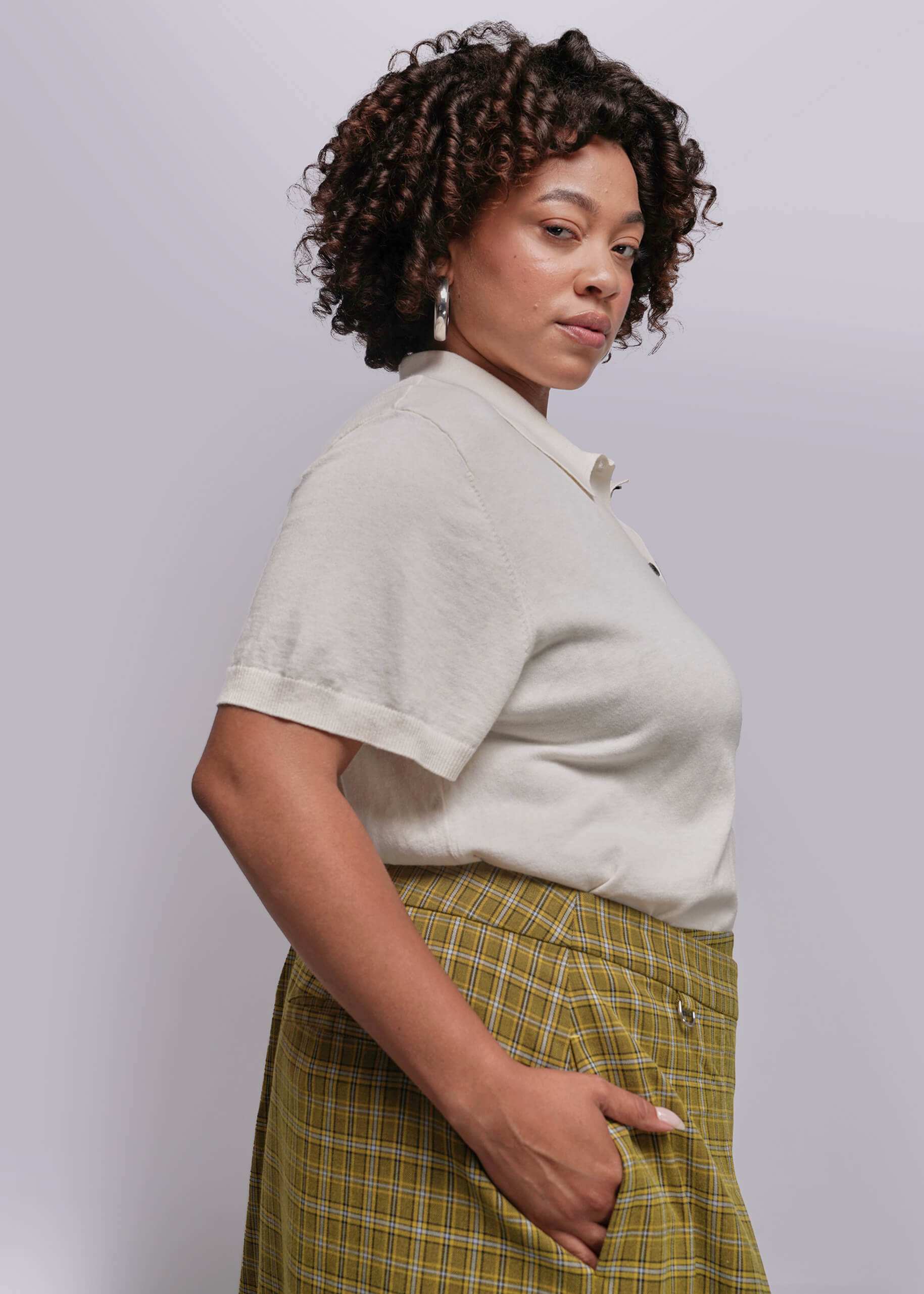 1X=Woman wearing a light-colored shirt and green plaid skirt against a plain background