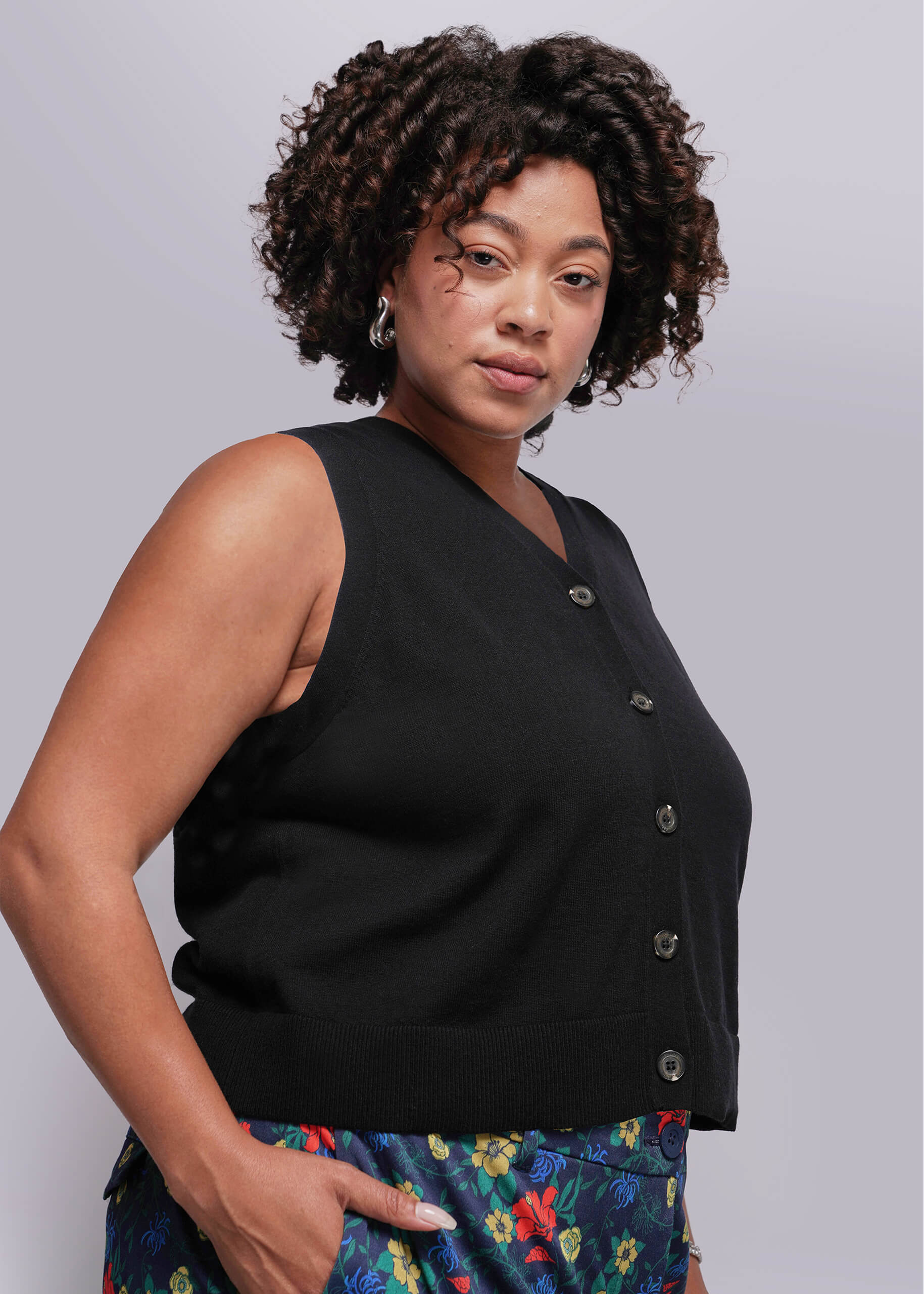 1X=Woman wearing a black sleeveless sweater vest top with buttons against a plain background