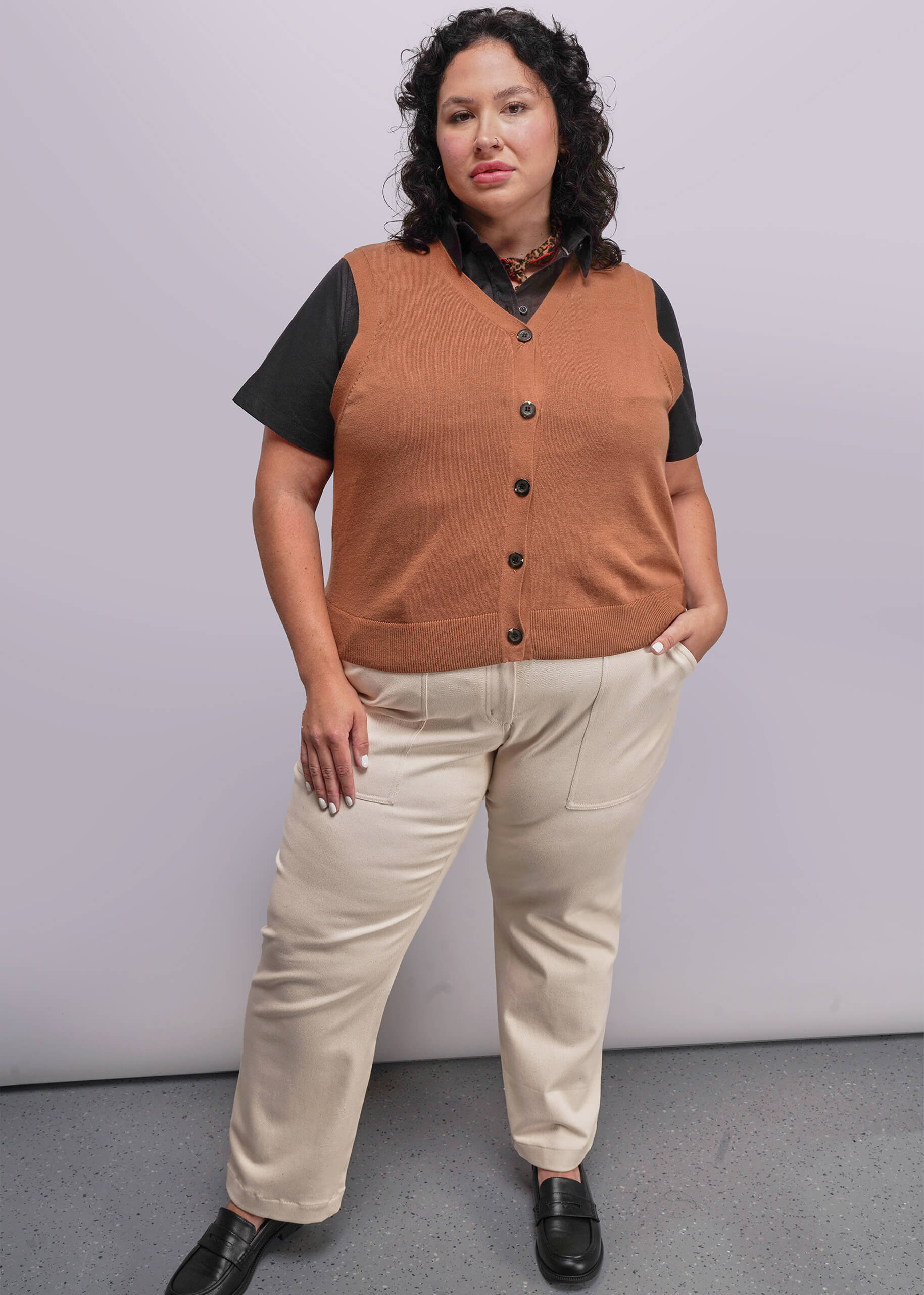 1X=Person wearing a brown vest over a black shirt with beige pants against a plain background