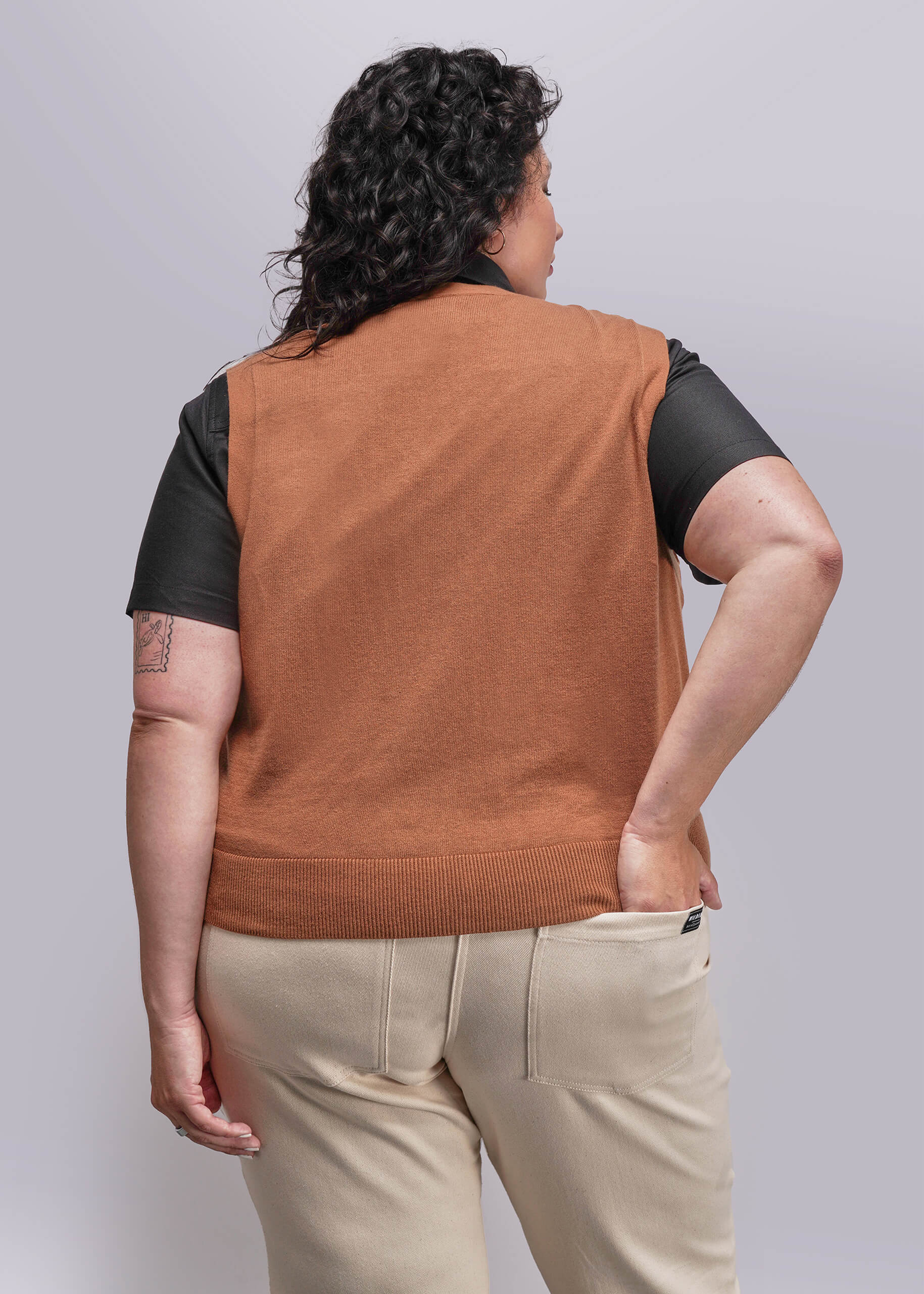 1X=Person wearing a brown vest over a black shirt with beige pants on a plain background