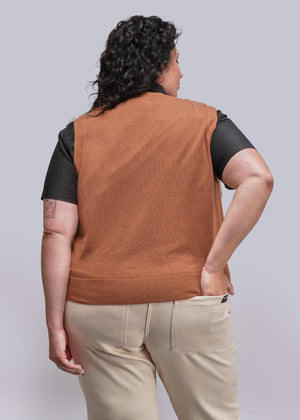 1X=Person wearing a brown vest over a black shirt with beige pants on a plain background