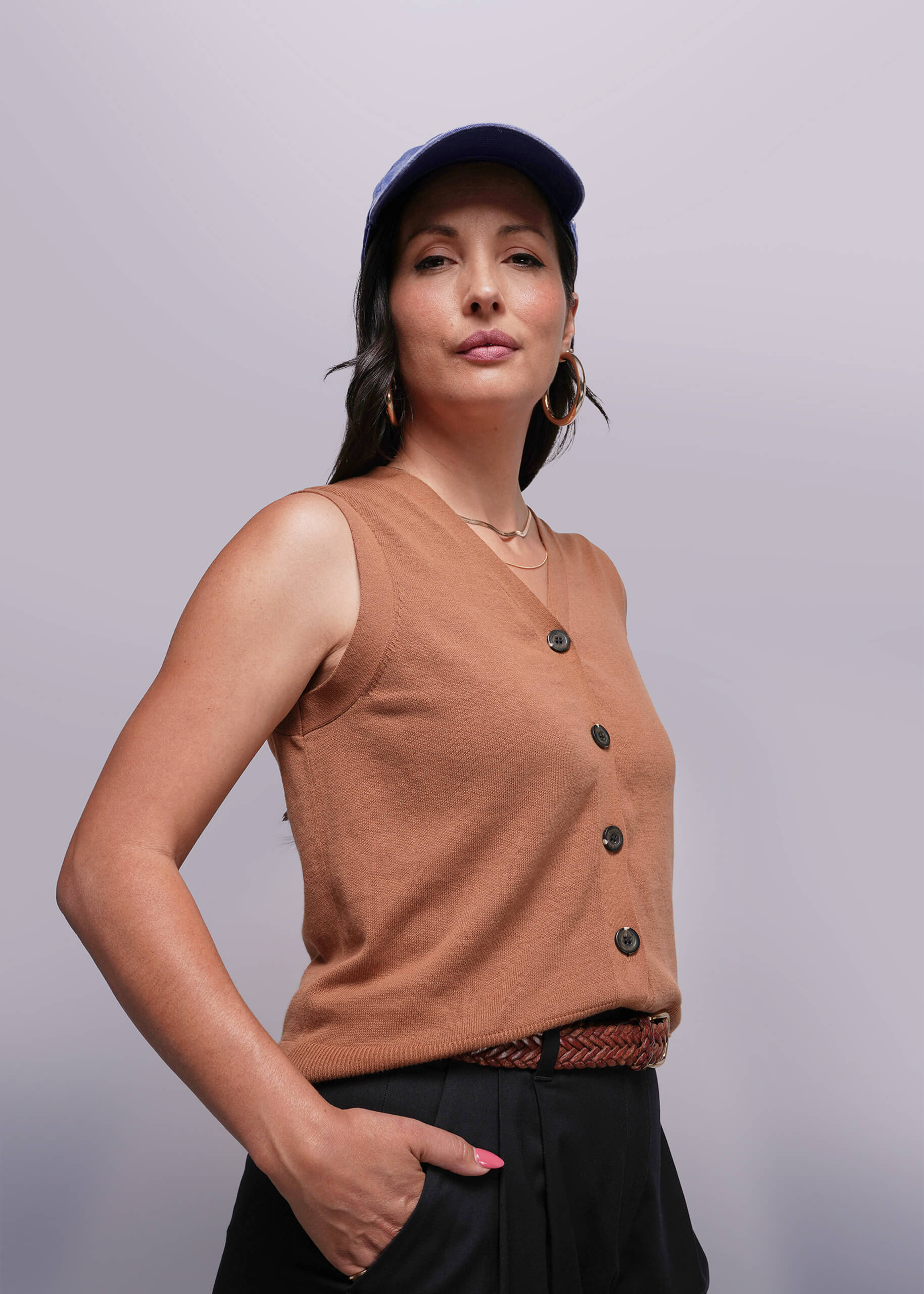 S=Woman wearing a brown sleeveless sweater vest with buttons and a blue cap against a plain background