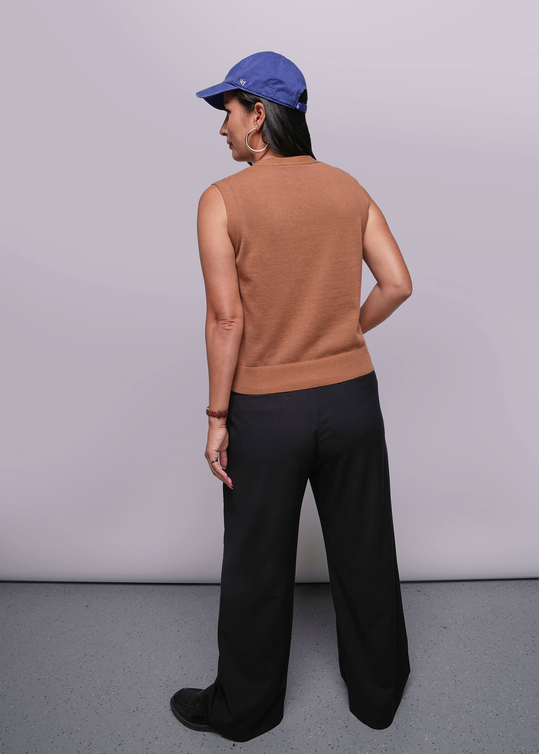 S=Person wearing a brown sleeveless top and black pants with a blue cap, standing against a plain background.