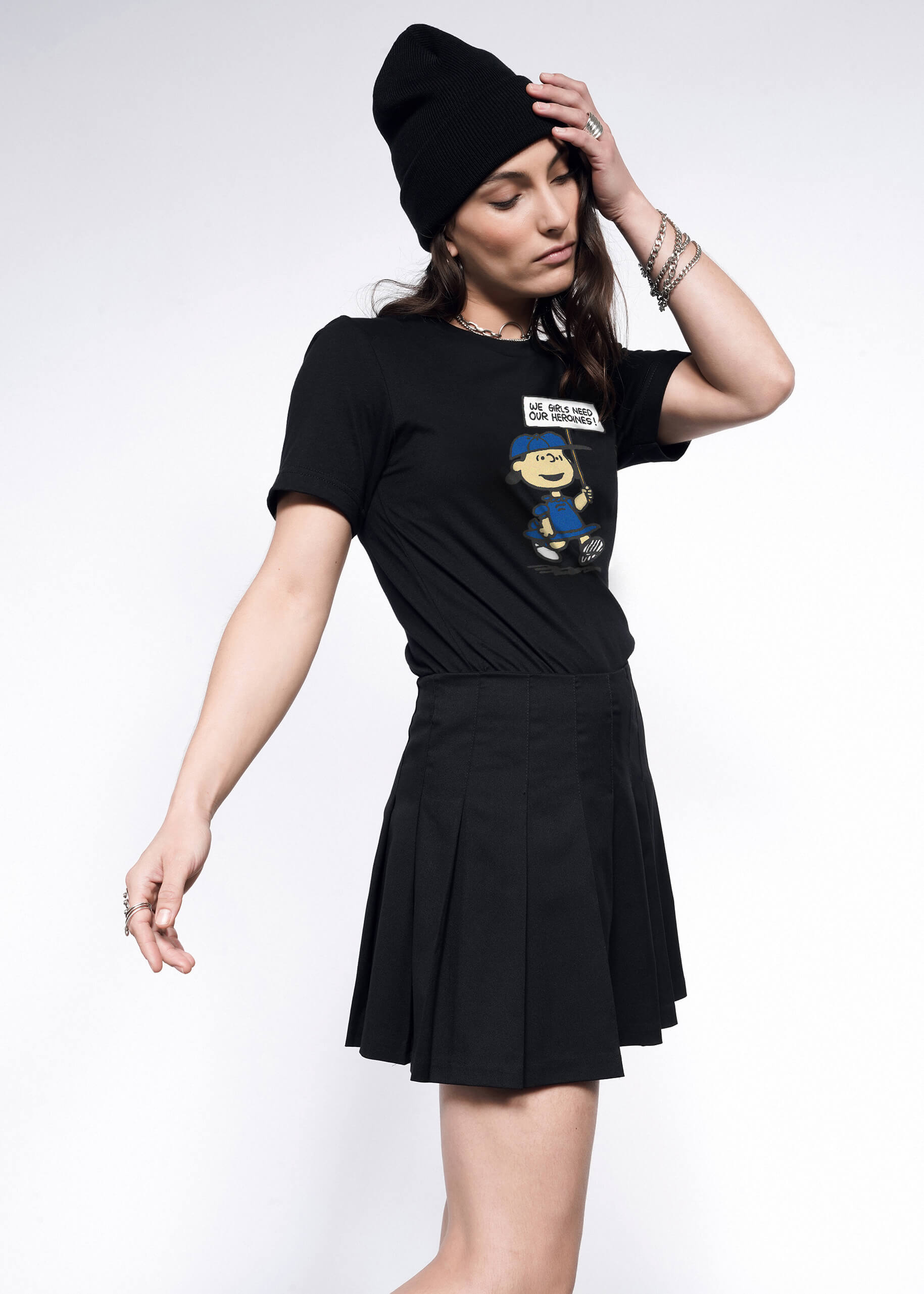 S= Side view of person with long brown hair wearing black beanie, black skirt, and The Peanuts x Wildfang Heroines Graphic Tee, which is a black tee that features a graphic of Lucy with a sign that says "We girls need our heroines"