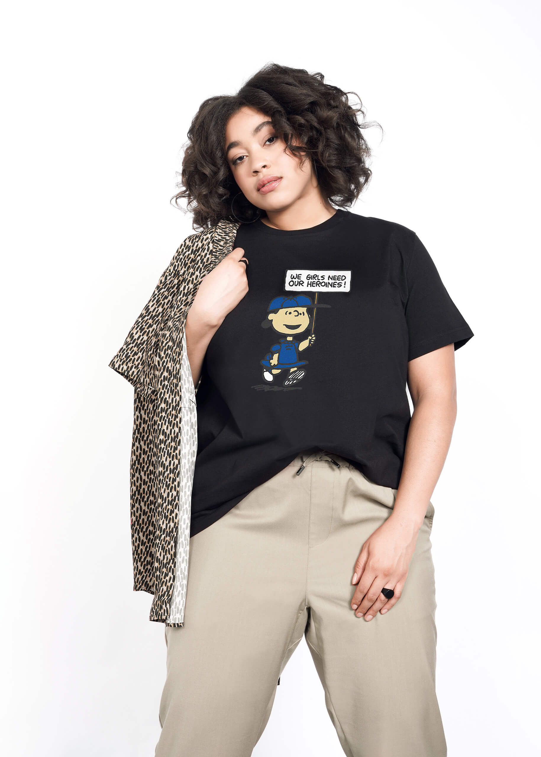 1X=Person with short curly brown hair wears tan trousers and The Peanuts x Wildfang Heroines Graphic Tee, which is a black tee that features a graphic of Lucy with a sign that says "We girls need our heroines"