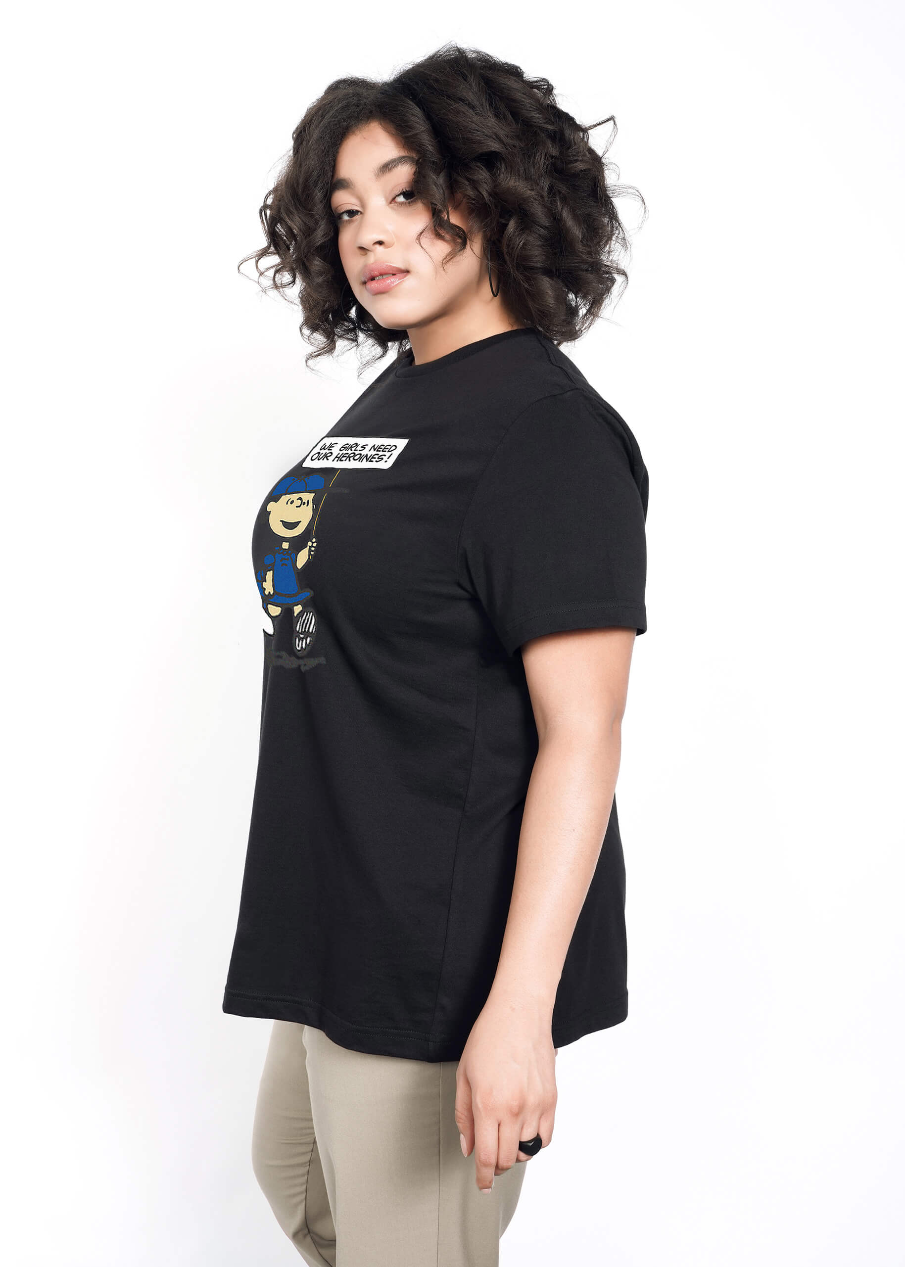1X= Side view of person with short curly brown hair wearing tan trousers and The Peanuts x Wildfang Heroines Graphic Tee, which is a black tee that features a graphic of Lucy with a sign that says "We girls need our heroines"