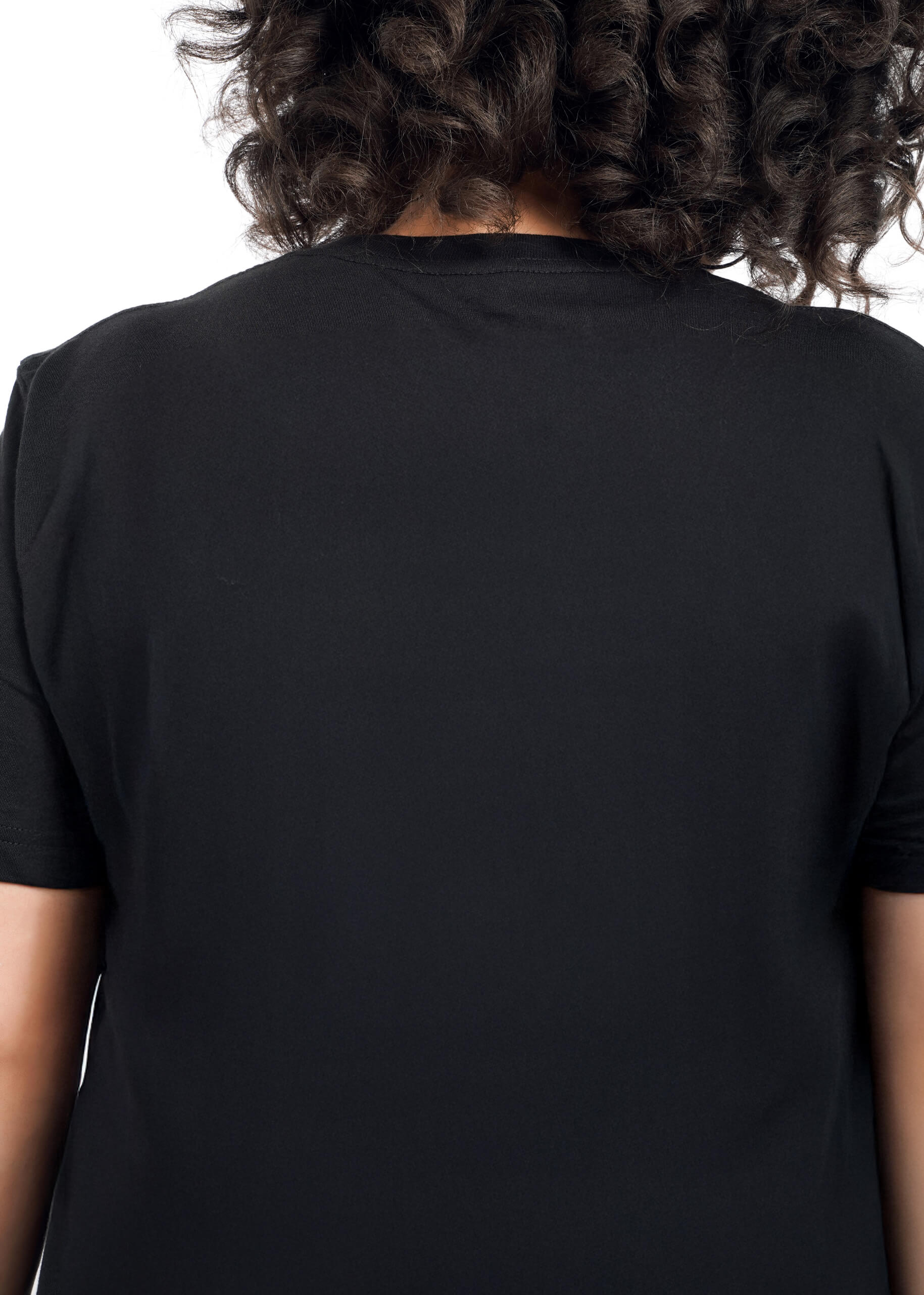 1X=Close up, back view of person with short curly brown hair wearing The Peanuts x Wildfang Heroines Graphic Tee, which is a black tee that features a graphic of Lucy with a sign that says "We girls need our heroines" Graphic is not in view in this image as this shows the back of the shirt.