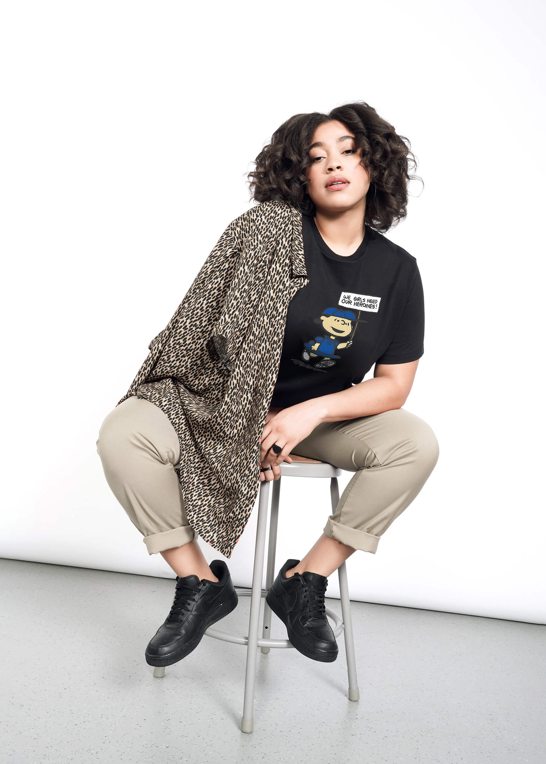 1X=Person with short curly brown hair wears tan trousers, black sneakers, and The Peanuts x Wildfang Heroines Graphic Tee, which is a black tee that features a graphic of Lucy with a sign that says "We girls need our heroines" They have a leopard printed jacket over their shoulder and sit on a stool.